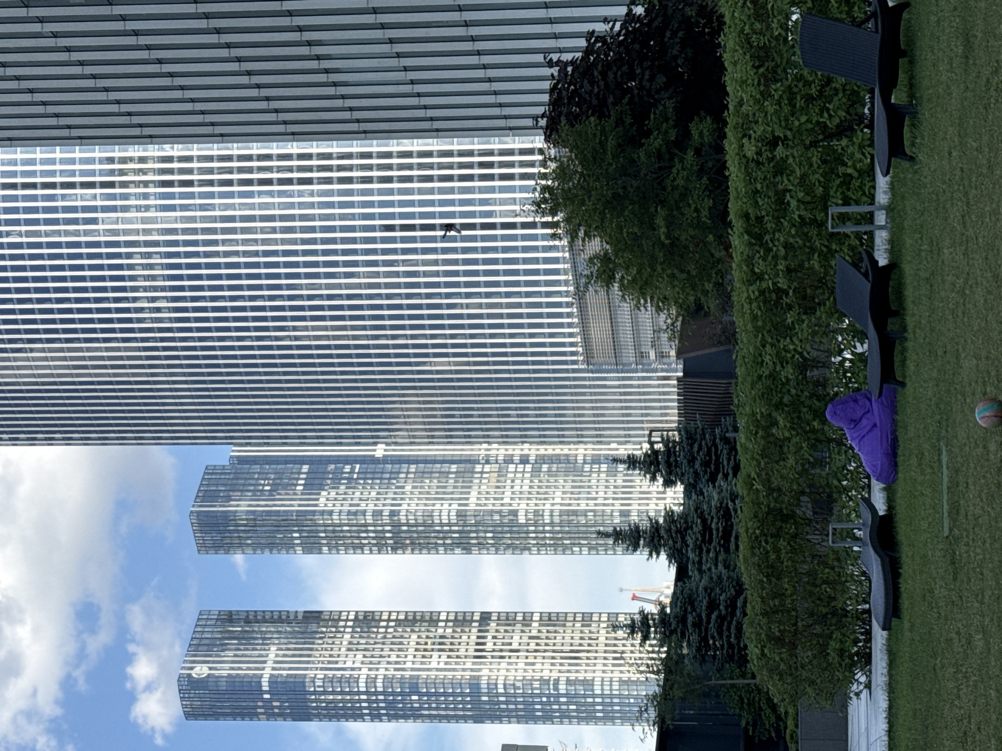 This daytime photograph captures a harmonious blend of modern urban architecture and a serene green leisure space in Moscow, Russia.
On the left side of the frame (when upright), several towering, contemporary skyscrapers dominate the background. Their sleek, reflective glass facades create intricate patterns of horizontal and vertical lines, mirroring the bright blue sky and scattered white clouds. The reflections give the buildings a dynamic and luminous appearance. A dark bird is visible in flight against the reflective surface of one of these skyscrapers. In the distance, between the buildings and foliage, a red and white construction crane can be faintly seen, indicating ongoing development.
The right side of the image (foreground when upright) features a well-maintained grassy lawn. Placed on the grass are several dark, possibly black or charcoal gray, outdoor lounge chairs, suggesting an area for relaxation. A vibrant purple garment is casually draped over one of these chairs. Nearby, a multi-colored ball, resembling a basketball or volleyball, rests on the grass. A dense line of green shrubs acts as a natural border, separating the lawn from the background trees and structures.
The weather is bright and clear with a blue sky and white clouds. The absence of people in the scene creates a sense of peaceful tranquility, highlighting a quiet urban oasis amidst the bustling city environment. No discernible text is clearly visible, apart from a faint, unidentifiable logo on one of the skyscrapers.