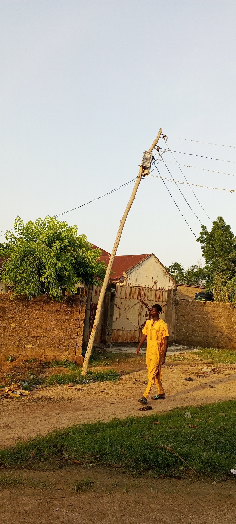 A man in a yellow traditional outfit walks on a dirt road in Jalingo, Nigeria. A slanted utility pole, with exposed wires and a meter box, stands prominently in the background. Behind the pole is a red-roofed building and a large, weathered metal gate. The environment is rural, with a low brick wall on the left side of the road and sparse vegetation. The sky is clear and light.