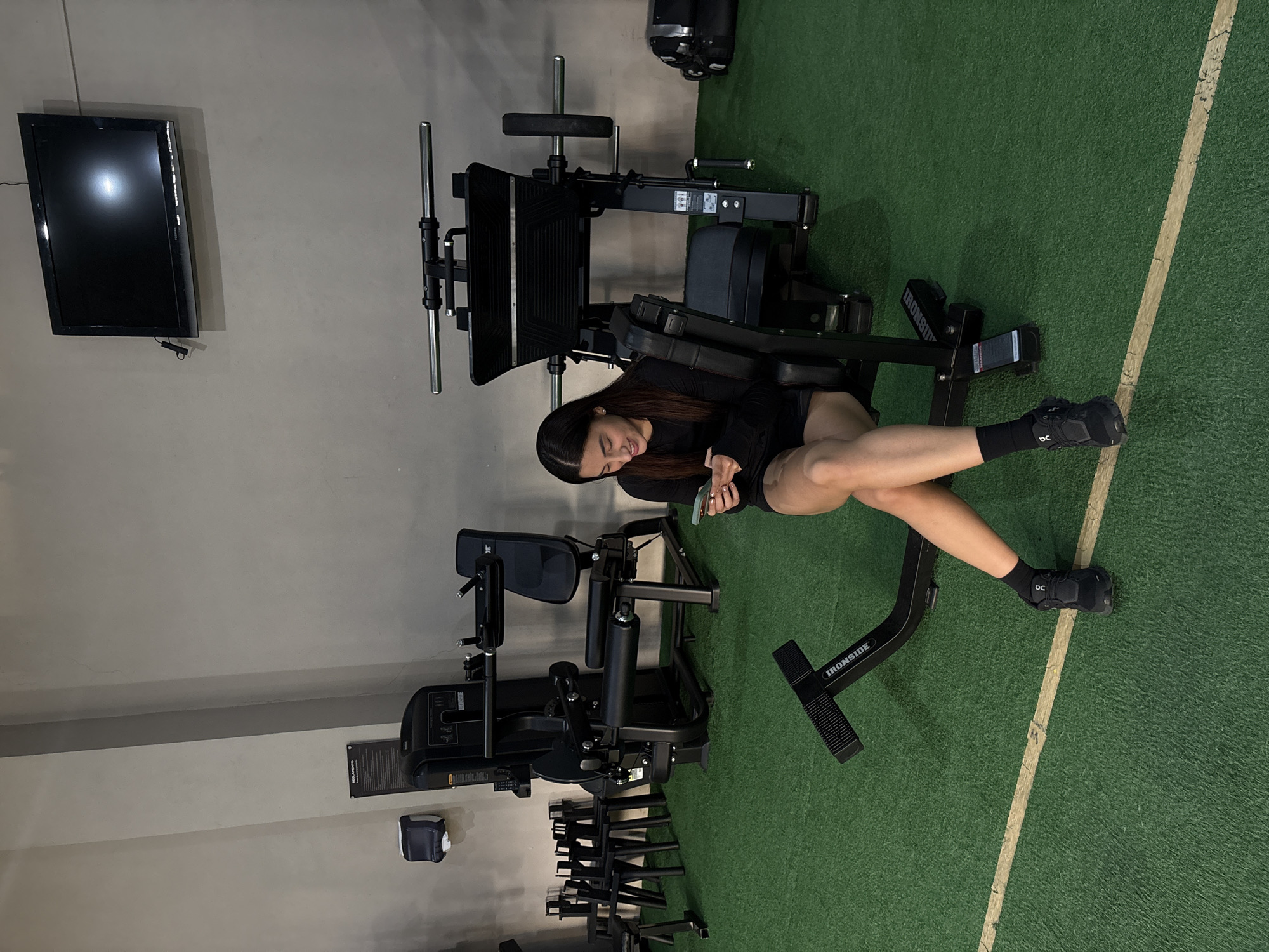 A woman in a black long-sleeved shirt and black shorts is sitting on an Iron Side fitness machine in a gym, looking at her phone. She's wearing black socks and black shoes. The gym has a green turf floor with a white line running across it. There's a TV mounted on the wall, along with various other gym equipment, including a leg press machine, and a weight rack. The location is San Bernardino Tlaxcalancingo, Mexico.