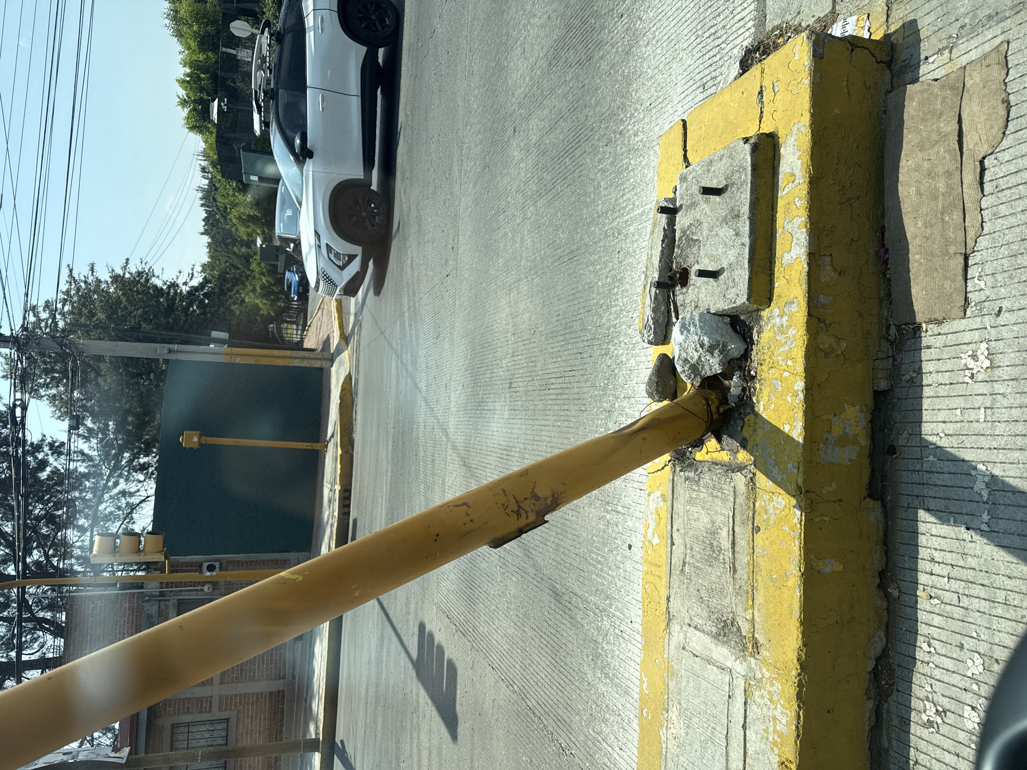 The image shows a yellow metal traffic barrier extended across a street in what appears to be a residential area. The barrier is attached to a yellow concrete support, which is chipped and peeling. Small rocks and debris are clustered around the base of the barrier support. In the background, parked vehicles (including a white SUV) are visible on the opposite side of the barrier. Wires and trees are visible in the distance. Based on the background and architectural details, it is plausible that the image was taken in Puebla, Mexico.