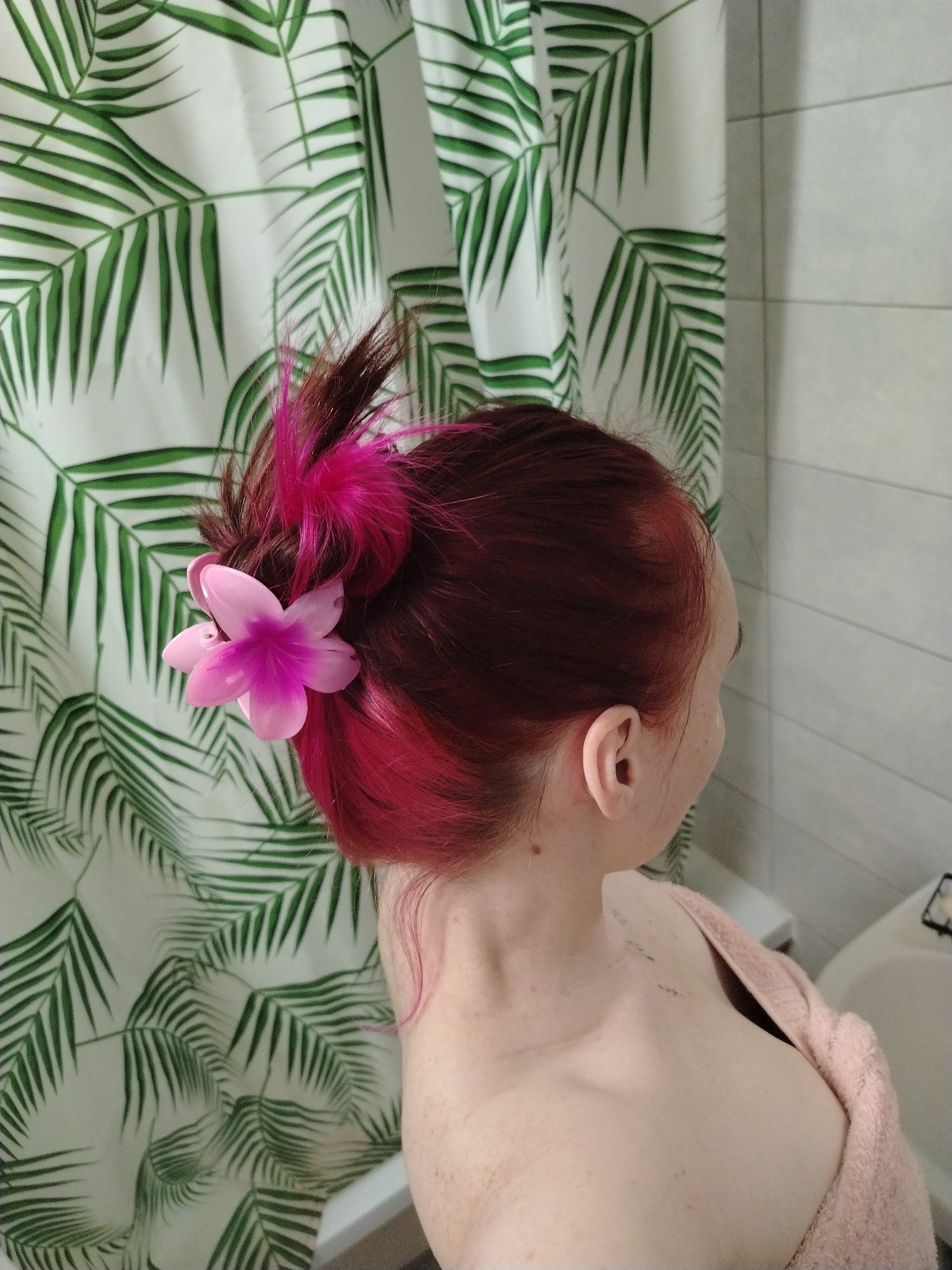 A woman with auburn hair styled in a bun with pink accents and a pink flower clip stands in a bathroom. Her right ear and shoulder are visible. She is partially covered by a peach-colored towel. Behind her is a shower curtain with a green and white palm leaf print and a tiled wall. A portion of a white bathtub is also visible. The location is not evident from the image.