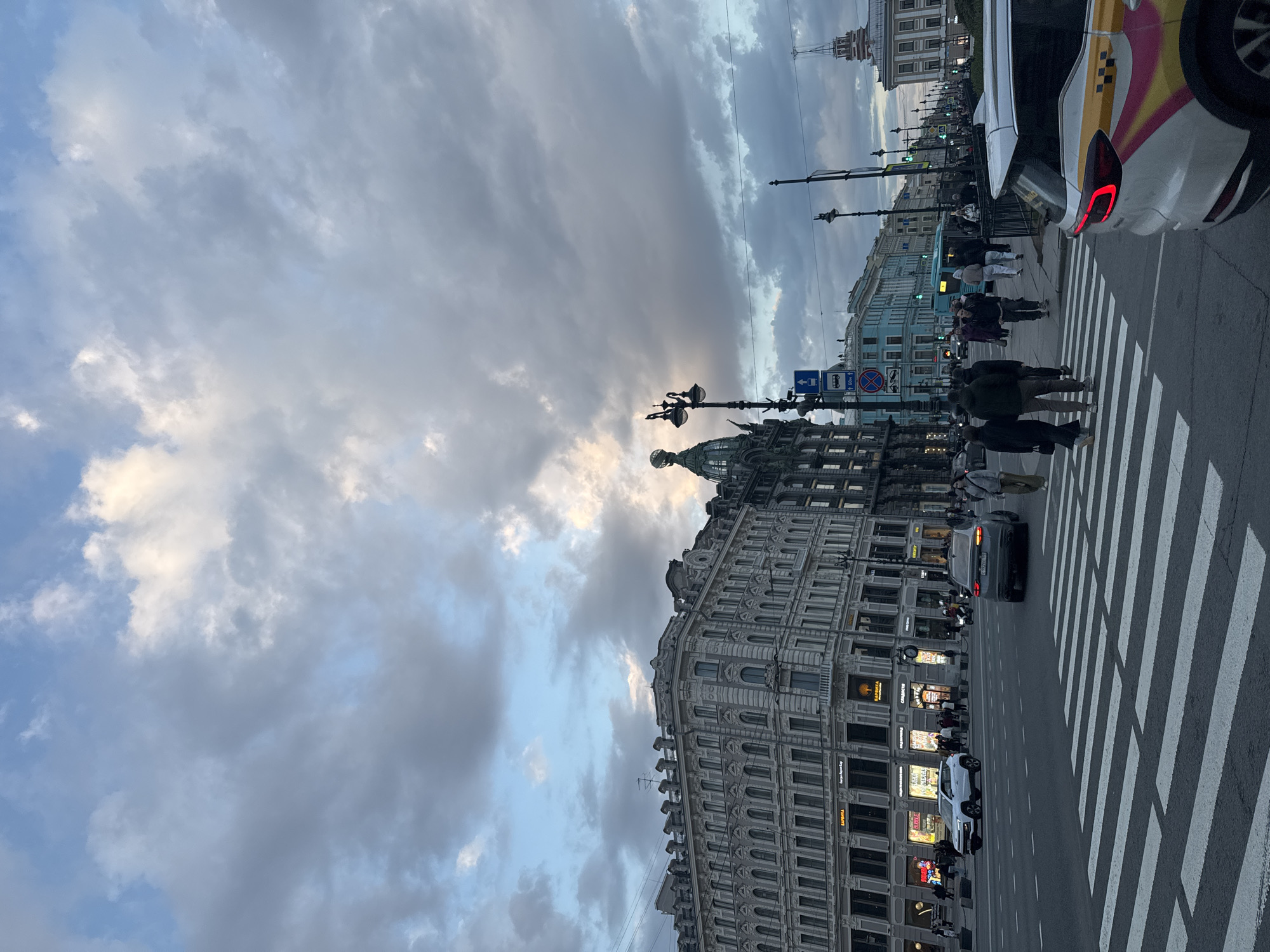 This media file captures a bustling street scene in Saint Petersburg, Russia, likely during the late afternoon or early evening under an overcast sky. The image is taken from street level, looking down a wide city thoroughfare flanked by impressive architecture.
The sky is dominated by thick, grey and white clouds, with patches of brighter blue showing through, indicating a break in the cloud cover or the diffused light of a setting sun. The overall lighting is soft, yet some ground-floor building lights are illuminated.
On the left side of the street, a grand, multi-story building with ornate, classical architecture stands prominently. It features intricate detailing, columns, and arched windows. A distinctive dome or cupola is visible at the top of an adjacent structure. Further down the street, other buildings, including one with a lighter blue facade, line the sidewalk.
The foreground and mid-ground are occupied by a wide, paved street with a clear, white zebra crossing. Several pedestrians are visible, some actively crossing the street on the zebra crossing, while others walk along the sidewalks. They appear to be dressed in everyday attire.
Vehicles are present on the street, including a white taxi with distinctive yellow and pink markings on its side, stopped near the crosswalk with its red taillight illuminated. Another dark-colored car is further down the road.
Street furniture includes tall, ornate black lampposts and various traffic signs. Visible signs include a blue rectangular sign with a white downward arrow and a bus symbol, indicating a bus stop or lane, and a red circular sign with a diagonal line, signifying no stopping or parking. Another blue sign with a left arrow and bus symbol includes "80м," likely indicating a distance of 80 meters. On building facades, some Cyrillic text is visible, including "БАРБЕРА" (BARBERA) and possibly "ГОСТИНЫЙ ДВОР" (Gostiny Dvor), which is a well-known location in Saint Petersburg.
