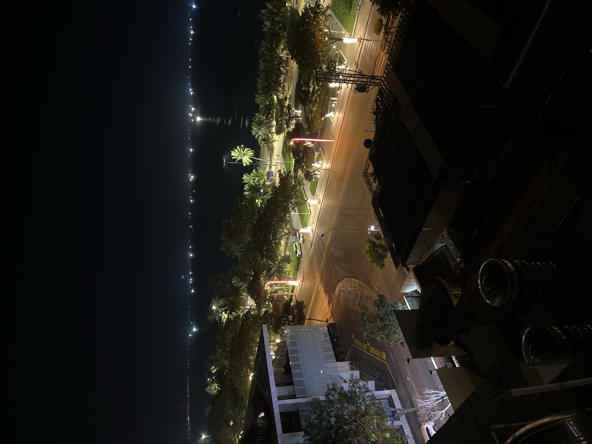 This is a night-time, high-angle shot of a coastal city street in Nha Trang, Vietnam, overlooking the ocean. In the foreground, dark rooftops and large water tanks are visible. The midground features a modern building with a white facade and glass elements, adjacent to a street with visible lane markings and sidewalks. Palm trees and other vegetation line the street, illuminated by streetlights and decorative lighting. Cars are parked on the side of the road, and there is a pedestrian crossing. The background shows the dark ocean stretching to the horizon, dotted with numerous lights from boats or ships, and the distant city lights along the coastline. The overall impression is of a well-lit urban area at night by the sea.