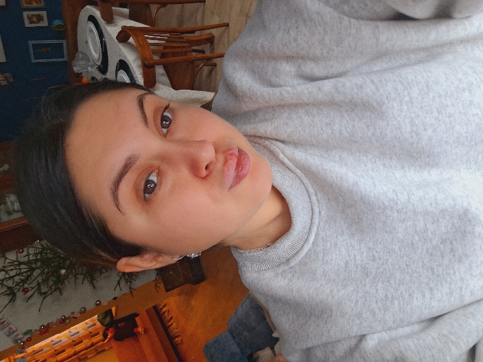 A female individual, appearing to be an adult in her 20s or 30s, is depicted in a selfie, positioned centrally and looking directly at the camera with pursed lips. She has dark hair, brown eyes, and is wearing a light grey crew-neck sweatshirt and a silver chain necklace. Small, silver-colored dangling earrings are visible. The scene is an indoor residential interior in Lezhnevo, Russia.
To the subject's left, a decorated Christmas tree with ornaments stands near a window or doorway adorned with string lights. A wooden bench or low table is visible beneath the window/doorway, with a dark blanket or pillow on the floor nearby.
To the subject's right, a dining area is visible, featuring a wooden table covered with a white tablecloth. Two place settings are present, each with a white plate atop a dark placemat. A clear glass pitcher or carafe is also on the table. Two wooden chairs with curved backs are positioned around the table. The wall in this section is dark blue and features several framed pictures or decorative items. The lighting in the room appears warm.