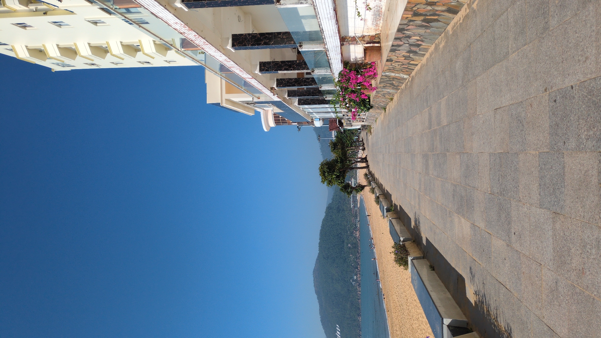 The image displays a sunny coastal scene in South Quy Nhon Ward, Vietnam, featuring a tiled promenade running parallel to a sandy beach and the ocean. The foreground is dominated by a wide pedestrian walkway paved with rectangular grey stone tiles, extending into the distance. To the left of the walkway, a low concrete wall with dark capping serves as a barrier, containing small green plants at intervals. Beyond this wall lies a wide, light brown sandy beach where several small, indistinct human figures are visible near the waterline. The calm, blue ocean stretches from the beach towards a distant, verdant mountain range, which features large white lettering on its left slope. To the right of the promenade, a series of buildings lines the path. The most prominent is a multi-story cream-colored structure with numerous balconies adorned with yellow accents, visible in the upper right quadrant. Adjacent to and below it are modern buildings characterized by glass balconies, dark marble-clad columns, and decorative stone mosaic paneling on their lower walls. Planters containing vibrant pink flowering plants are positioned along the base of these structures. The sky overhead is a clear, uninterrupted deep blue, and strong sunlight casts distinct shadows across the promenade from the buildings and trees.