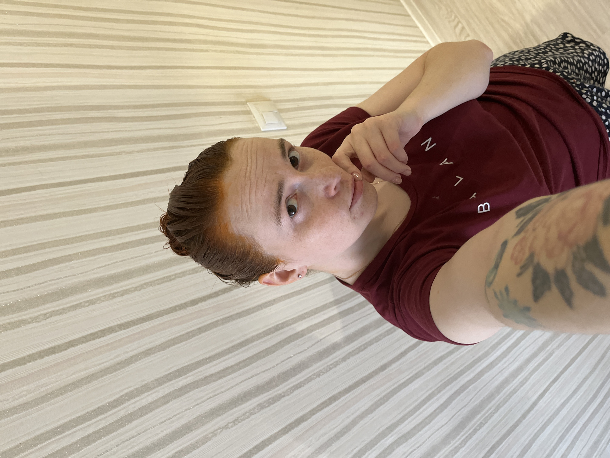 A pale-skinned person with red hair is pictured indoors. They are wearing a maroon t-shirt with white lettering that appears to spell out a name. The left arm, featuring a floral tattoo, is outstretched towards the camera. The person's right hand is resting on their face. The background features a horizontally striped wall. A light switch is visible in the top left corner of the image. Part of what appears to be a printed skirt or shorts is visible to the right of the frame. The person's gaze is directed towards the camera. This image was taken in the city of Chaikovsky, Russia.