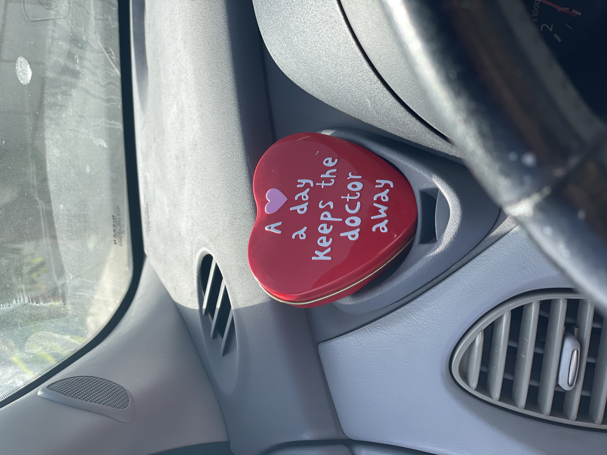 A red heart-shaped tin with white text reading "An apple a day keeps the doctor away" is placed in a car's dashboard vent. The tin has a small pink heart above the text. The dashboard is gray, and a portion of the steering wheel and speedometer are visible in the foreground. The scene takes place inside a vehicle.