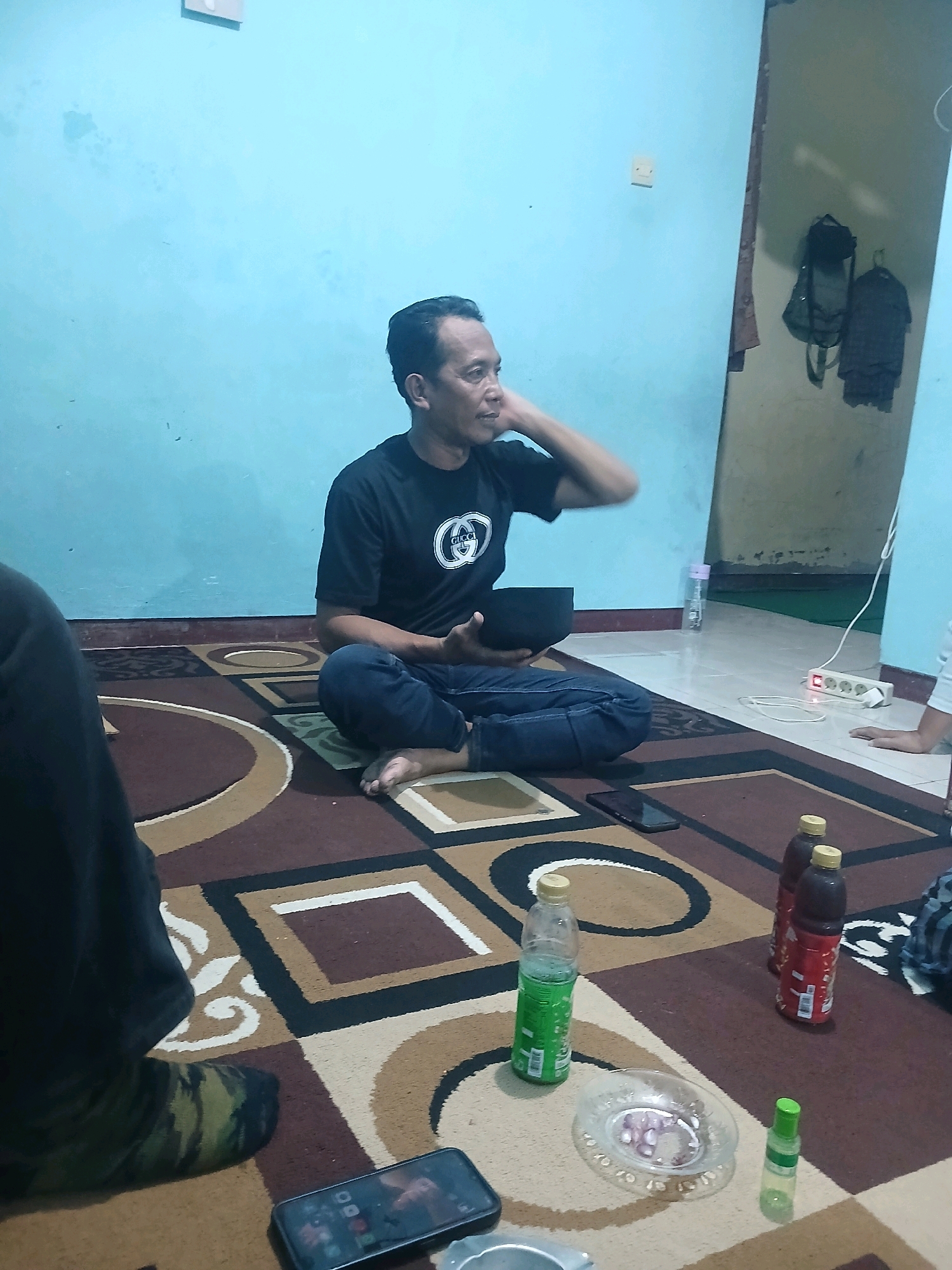 A man wearing a black t-shirt and dark jeans sits cross-legged on a carpeted floor. He is holding a dark bowl in his lap with his right hand and appears to be scratching his head with his left hand. The man's right foot is visible, with his toes pointing upwards. A patterned carpet covers the floor, with abstract shapes in brown, beige, and white. To the right of the man, on the carpet, are two bottles of dark liquid with red labels and yellow caps, a green plastic bottle of liquid, a small glass dish containing what appears to be sliced red onions, and a small green bottle. A smartphone lies on the carpet near the man's feet. In the background, a light blue wall is visible, with a white electrical outlet on the wall. To the right, a doorway leads to another room where backpacks and clothing are hanging on the wall. The scene appears to be indoors, likely in a residential setting in Banyuwangi, Indonesia.