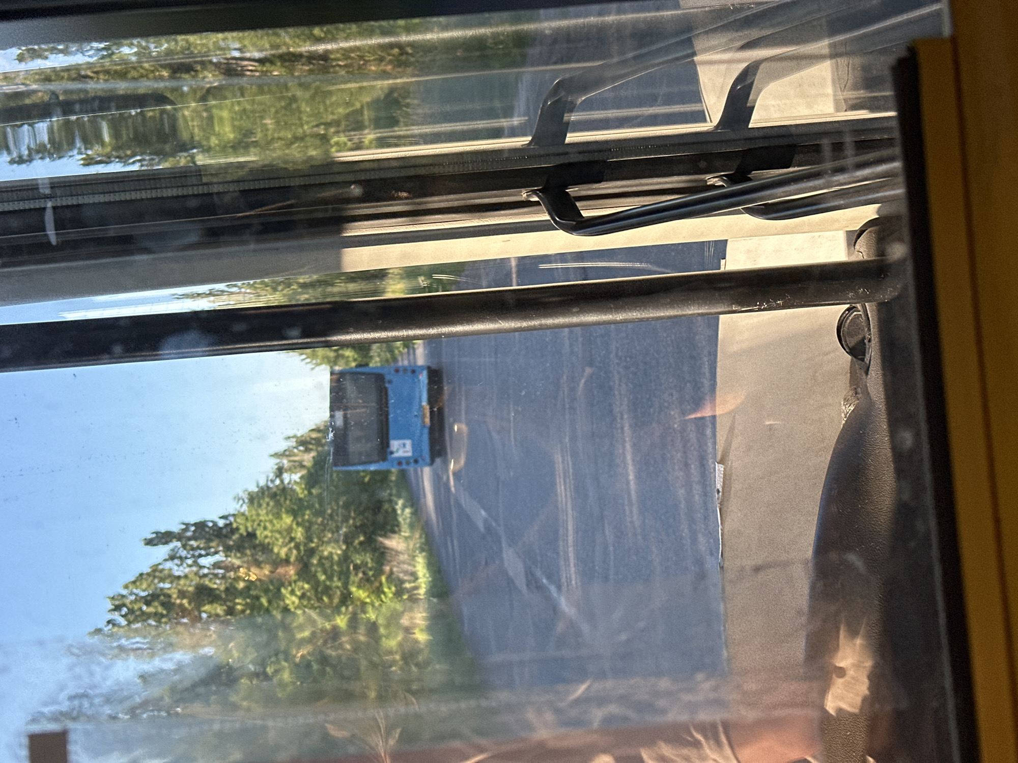 The image is a photograph taken through a window, possibly of a bus or train, looking outwards. The perspective is from inside, with the window frame and some interior elements, such as metal bars and possibly a seat, visible in the foreground and along the edges.
The main subject visible through the window is a blue bus driving on a road. The bus is seen from behind and at an angle. The road is paved and appears to be relatively smooth. On the left side of the road, there are trees and foliage, suggesting a natural or semi-rural setting. The sky is visible above the trees and appears to be a clear blue, indicating daylight. The lighting suggests it might be late afternoon or early morning, given the warm tones and long shadows cast by the sun, especially on the road.
Reflections are also present on the window, adding to the complexity of the scene. These reflections obscure some details and create a layered effect. The overall impression is of a journey in progress on a pleasant day. The location context provided is Dmytrivka, Ukraine. No visible text is present in the image.