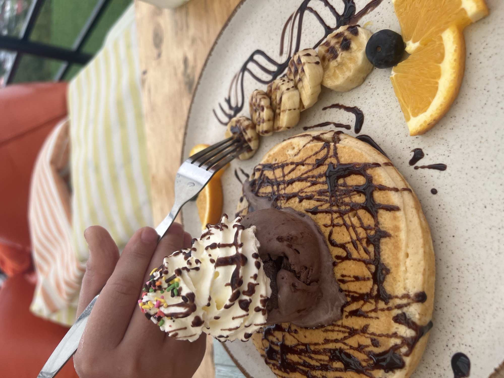 A close-up, overhead shot shows a dessert plate on a wooden table. The dessert consists of a large, round pancake topped with a scoop of chocolate ice cream, a swirl of whipped cream, and colorful sprinkles. Chocolate syrup is drizzled over the pancake and ice cream. To the right of the pancake are several slices of banana, also drizzled with chocolate syrup, and a slice of orange with a blueberry on top. A fork is being used to cut into the dessert. The background is blurred, showing an orange cushion and a striped fabric. This image was taken outdoors in Ko Samui District, Thailand.