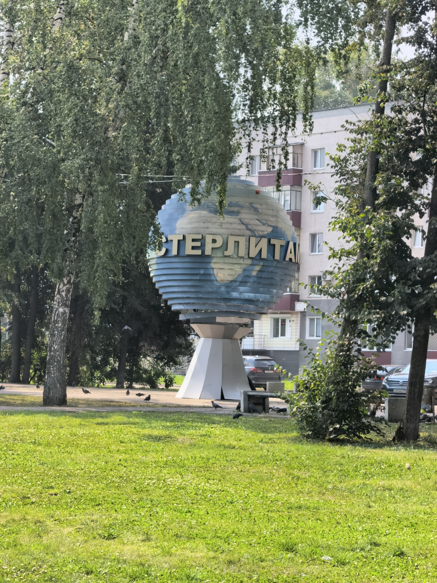 The image shows a large, blue and white globe sculpture with the word "СТЕРЛИТАМАК" written in yellow letters. The sculpture is located in the city of Sterlitamak, Russia. It is situated on a paved area in front of a light-colored, multi-story building. Trees with green foliage surround the sculpture, and a patch of well-maintained green grass is in the foreground. Several pigeons are scattered on the pavement and grass. Cars are parked near the building. The scene appears to be an outdoor public space on a sunny day.