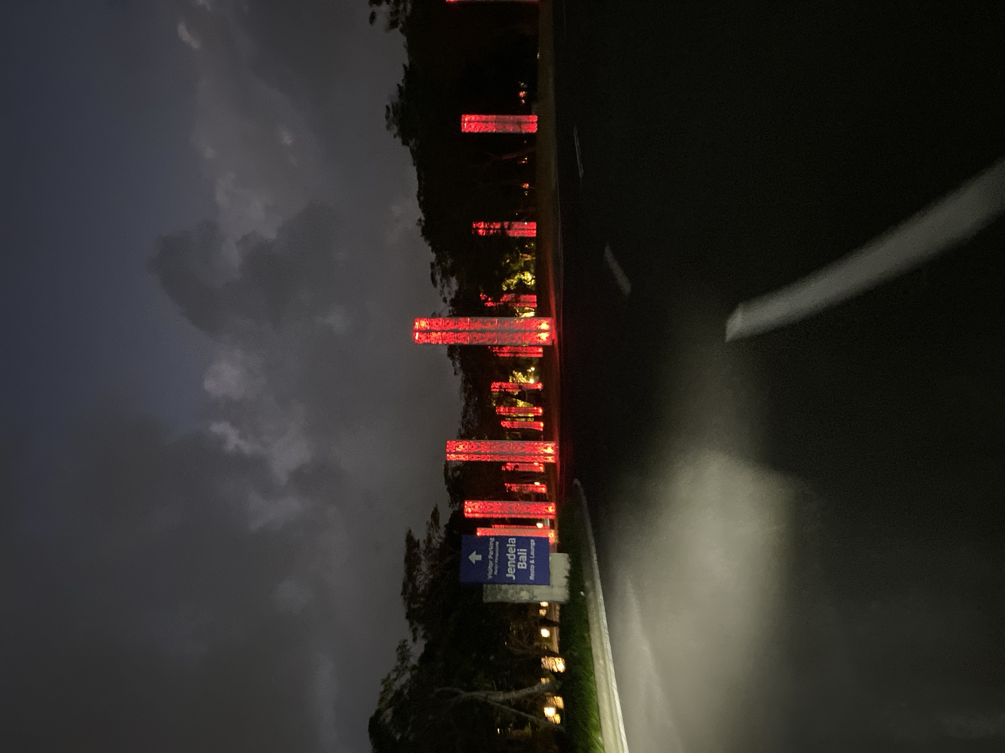 The image shows a road at night in Kuta Selatan, Indonesia, leading towards illuminated structures. Tall, rectangular pillars glow with bright red light, arranged in a line along the side of the road. To the left of the road, a blue sign with white text indicates "Visitor Parking" and "Jendala Bali Resto & Lounge." The sky above is dark with scattered clouds. The road surface is visible in the foreground, with a bright headlight beam illuminating a portion of it. Trees and other foliage form a dark silhouette against the background. The scene suggests an entrance to a venue or establishment that is decorated with prominent lighting for nighttime appeal.