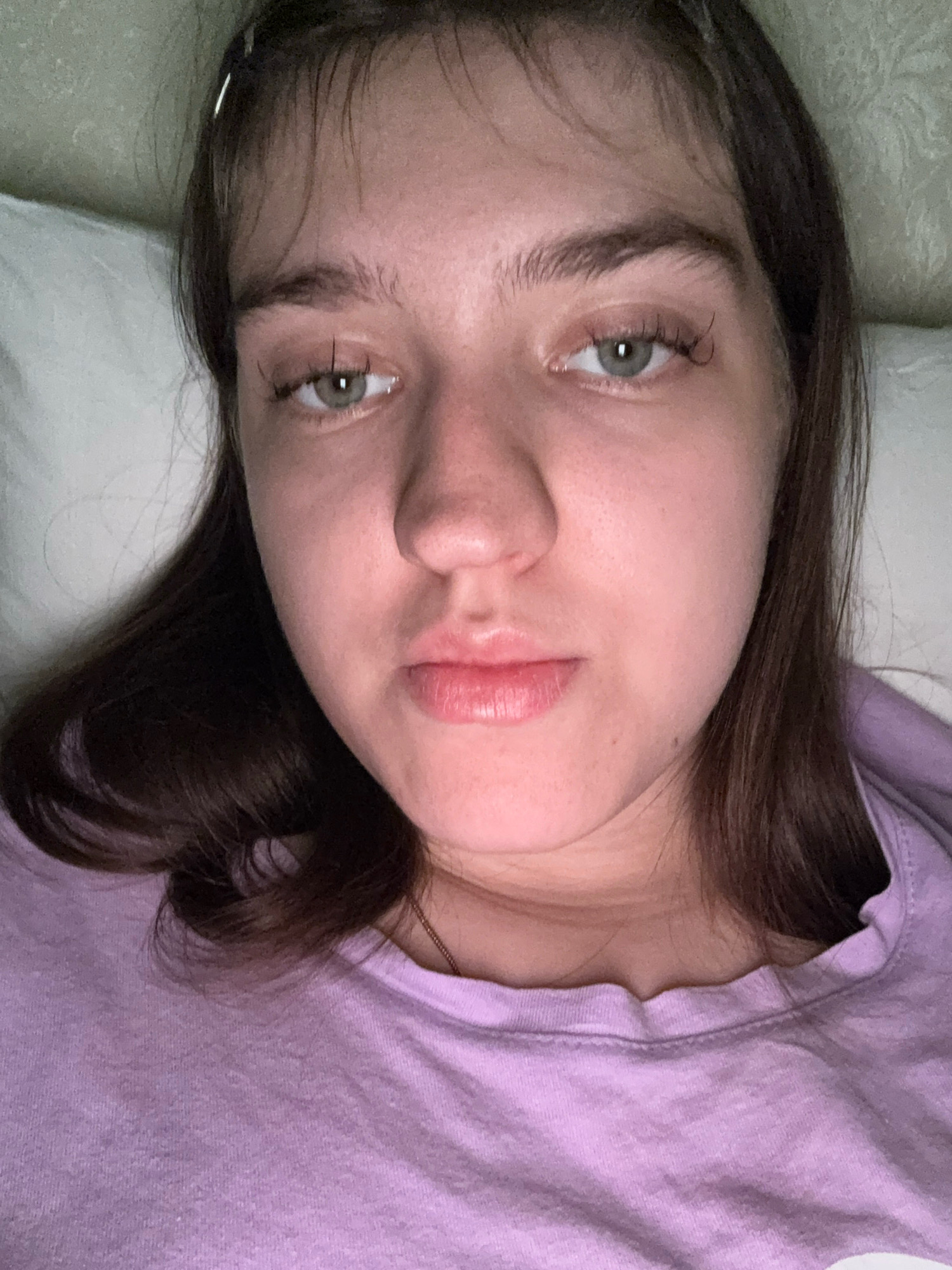 The image is a close-up, overhead shot of a young woman lying down. She has brown hair and green eyes and is wearing a purple t-shirt. The background appears to be a white pillow and a textured wall. The lighting is dim, suggesting an indoor setting, possibly a bedroom at night. The woman is looking directly at the camera with a neutral expression. There is no visible indication of the city of Zavolzhye, Russia, within the image itself.
