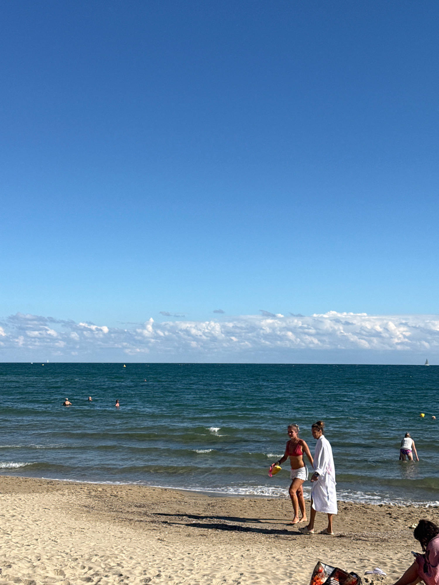A sunny day at a sandy beach in Sveti Vlas, Bulgaria, features several individuals engaged in seaside activities. The clear blue sky stretches above the tranquil, dark blue sea, where a few people are swimming. Closer to the shore, two women walk along the wet sand. One woman, wearing a red bikini top and white shorts, carries a yellow toy. The other, in a white bathrobe and light-colored shoes, walks beside her. In the background, another person stands in the shallow water. The sandy beach is speckled with debris, and a bag with what appears to be fruit is visible in the foreground. The scene is bathed in natural daylight.