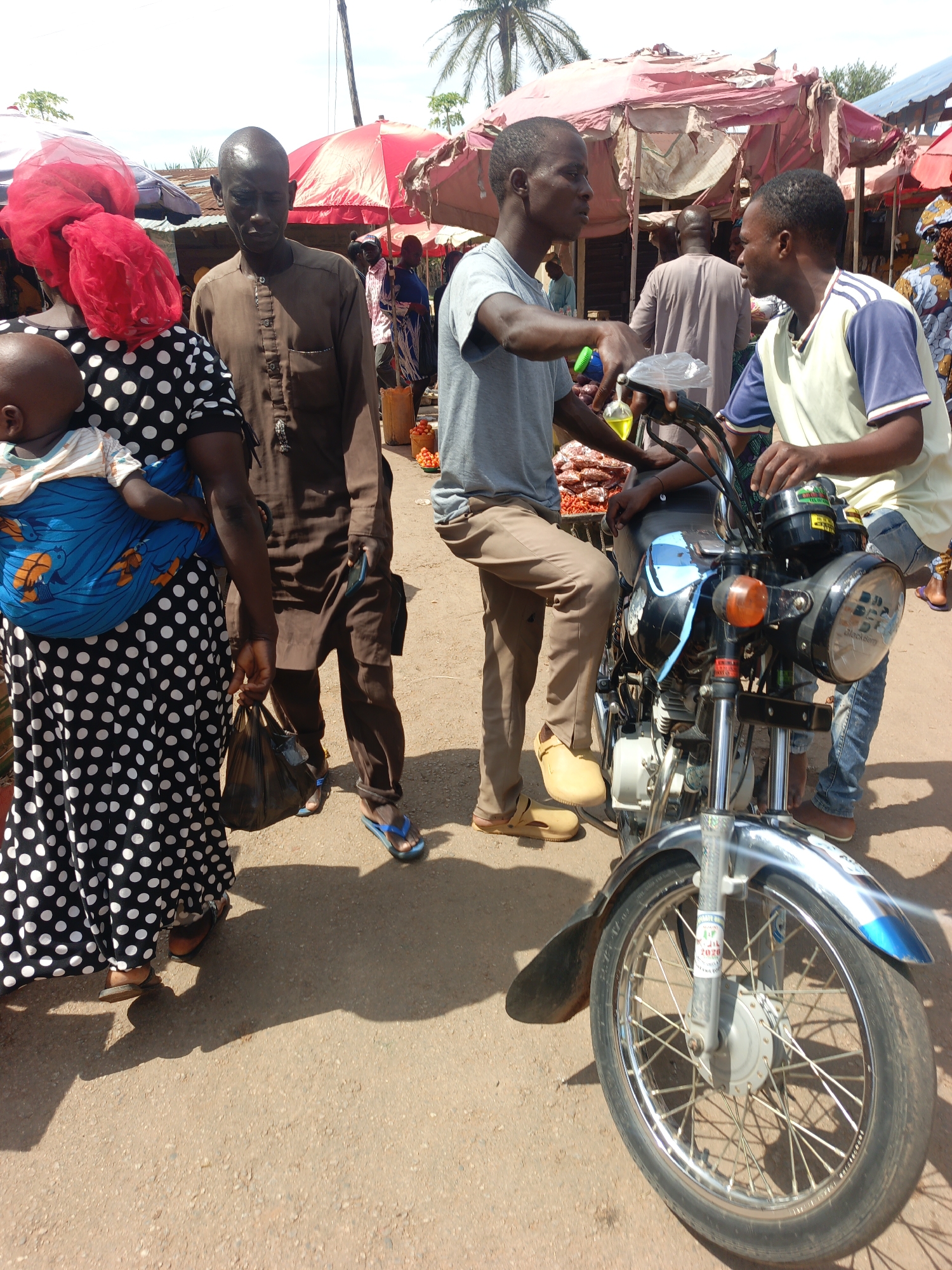 In Akwanga, Nigeria, a bustling outdoor market is captured during daylight. A woman in a black and white polka-dot dress and a red headscarf carries a baby in a blue sling. A man stands beside her, wearing a brown tunic and blue sandals. Two men are engaged in conversation near a black motorcycle with silver accents; one sits on the motorcycle, wearing a grey shirt and khaki pants, while the other stands, wearing a white and blue shirt and jeans. Market stalls with red and pink umbrellas are in the background. The ground is dirt and sand.