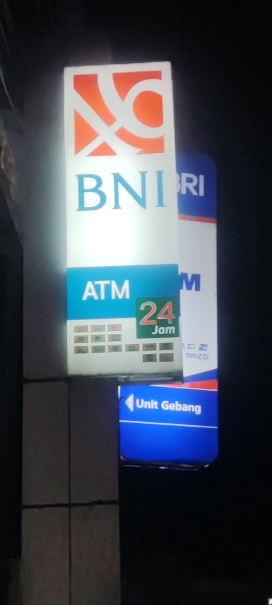A vertical, illuminated sign for BNI Bank is visible at night in Gebang, Indonesia. The sign features the BNI logo at the top in orange and white. Below the logo, the text "BNI" is displayed in large blue letters. Further down, "ATM" is written in white on a blue background. To the right of "ATM" is a green rectangle with "24 Jam" (24 Hours) written in orange and white. Below this, there are several small squares, possibly representing different payment card logos. The bottom portion of the sign is blue and contains white text and an arrow, indicating "Unit Gebang". To the right of the BNI sign, a portion of another sign is visible, displaying the text "BRI" and "BRIZZI". The surrounding environment is dark, indicating nighttime. The sign is mounted on what appears to be a building facade or structure.
