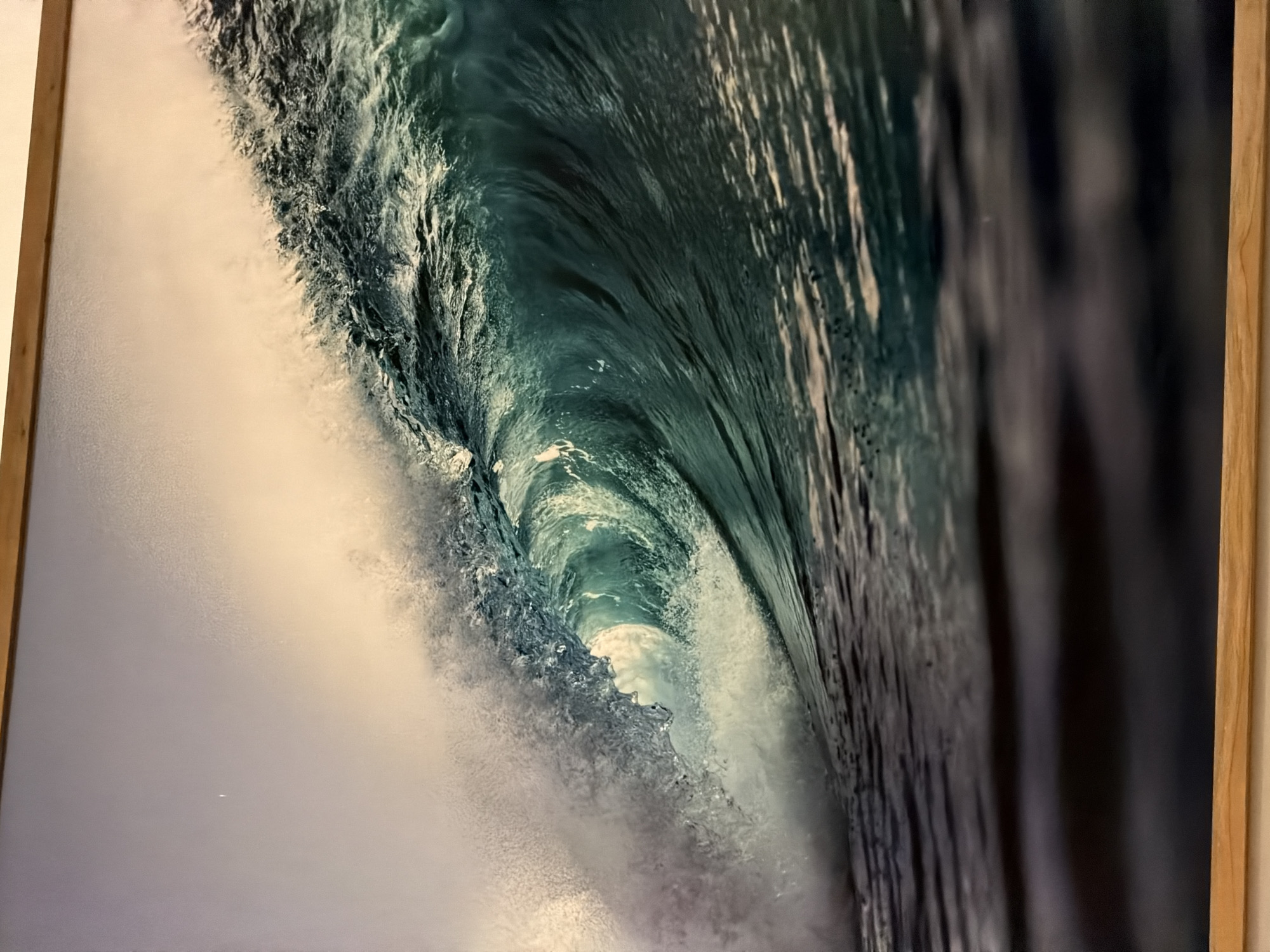 The image is a framed photograph of a breaking ocean wave. The wave is characterized by a turquoise inner curve, juxtaposed against frothy white water and dark, almost black, wave crests on the right side of the image. The photograph is likely taken from within the wave's barrel. Based on the wave characteristics, it is possible this image was taken in Kuta, Indonesia, a popular surfing destination known for its powerful waves, particularly during the wet season. There are no people visible. The focus is on the natural element. The photograph is contained within a light-wood frame.