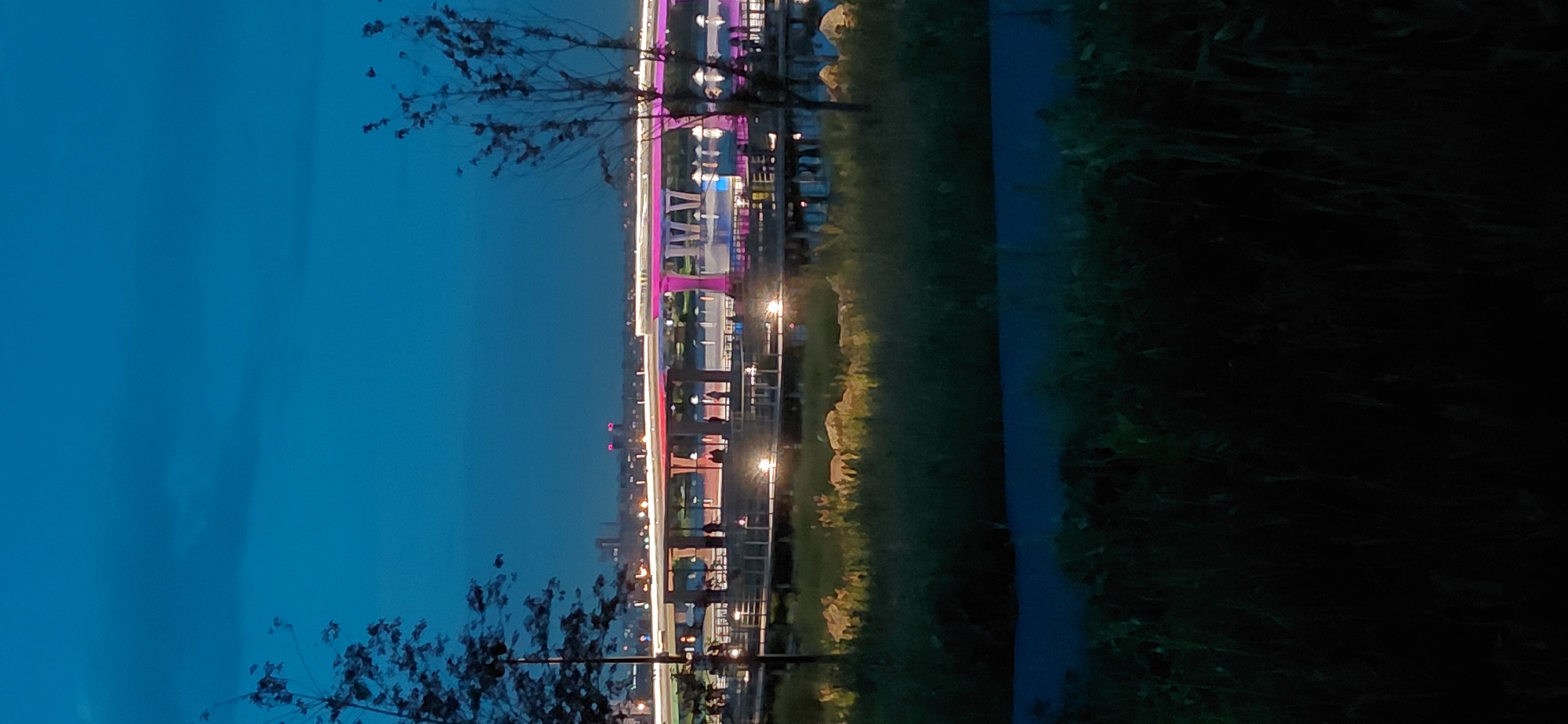 The image depicts a twilight scene with a broad, multi-lane bridge illuminated by white and pink lights. The bridge spans across a body of water, with a promenade or walkway visible beneath its deck, lined with glowing lights and populated by several silhouetted figures. The foreground features dark, out-of-focus vegetation, and a paved path is visible. In the distance, the city skyline is visible, characterized by a few tall buildings and other illuminated structures. The sky is a deep blue, indicative of evening. The scene captures a public space, likely a park or waterfront area, during dusk or early night. The location in Magnitogorsk, Russia, is suggested to be near the city's bridge.