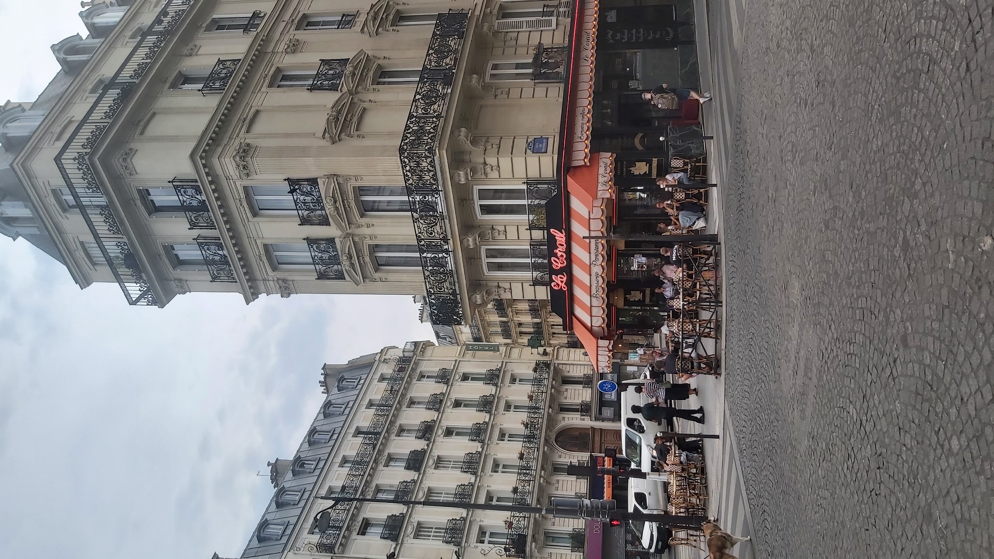 The image captures a street scene in Paris, France, with a focus on a corner building housing a brasserie named "Le Corail". The brasserie features an orange and white striped awning and outdoor seating where patrons are visible. The architecture of the building is typical of Parisian Haussmannian style, with ornate balconies and mansard roofs on the adjacent building. Several vehicles, including white vans, are parked or driving on the street, and cobblestone pavement is prominent. A dog is walking across a crosswalk in the foreground. The sky is overcast, suggesting a cloudy day.