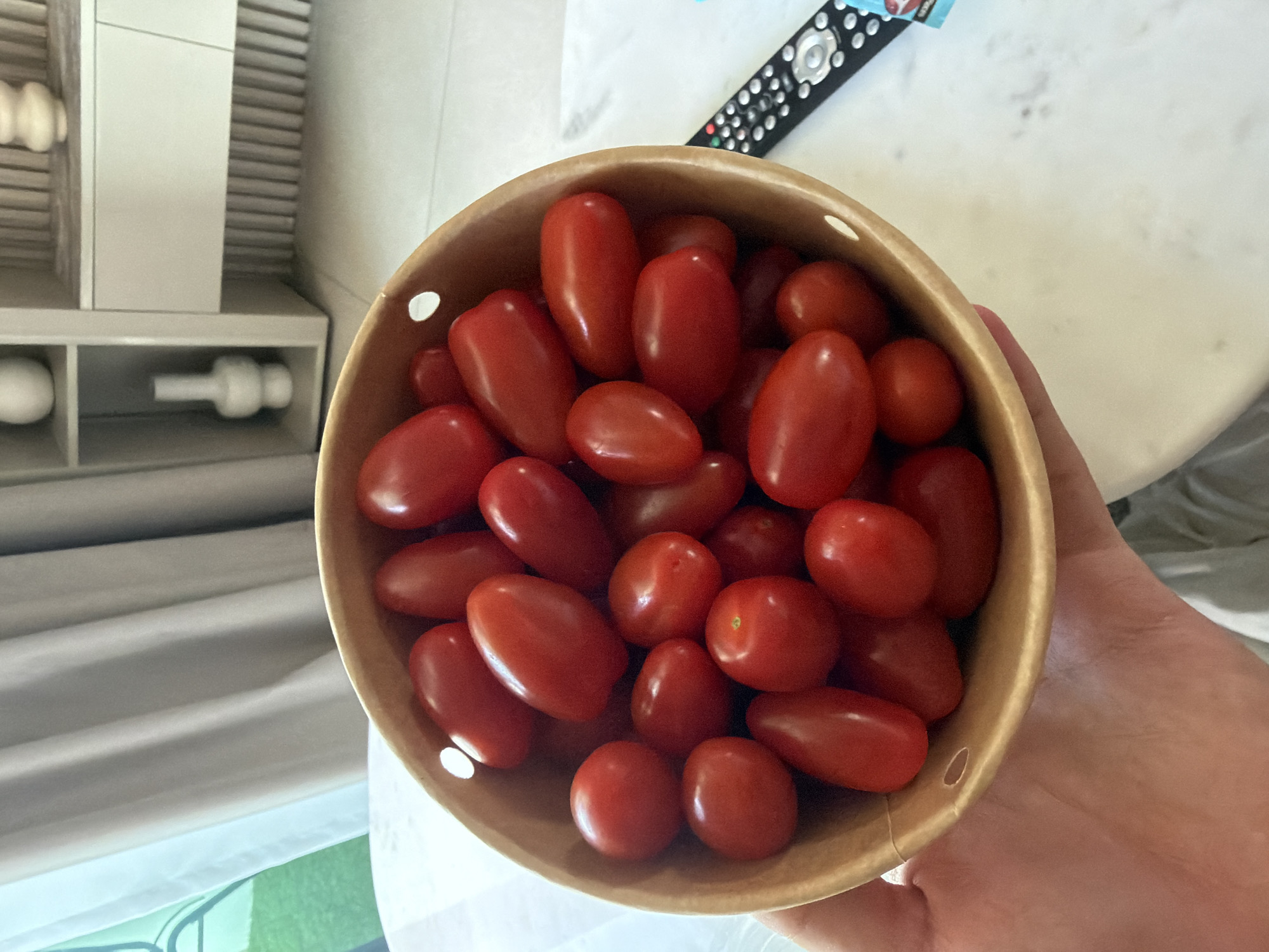 The image shows a close-up, top-down view of a tan-colored bowl filled with ripe, red plum tomatoes. The bowl is being held in a person's hand. The setting appears to be indoors, possibly a kitchen or dining area, as suggested by the presence of a white cabinet unit with white cylindrical objects on the left side of the frame. In the background, there's a remote control lying on a marble or white surface. There is no information in the image to indicate the location in Dubai, United Arab Emirates.