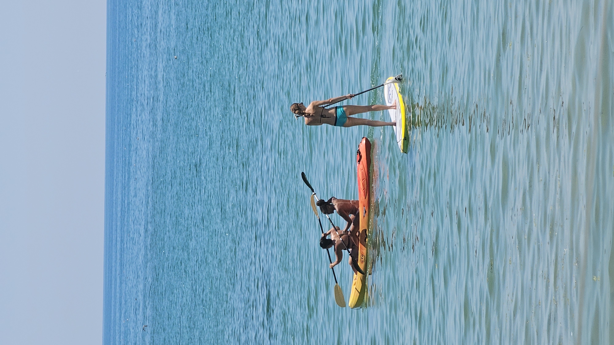 The image depicts a bright, sunny day at the Black Sea coast in Varna, Bulgaria. In the foreground, a person stands on a yellow and white paddleboard, holding a paddle. To the left, a two-person kayak is occupied by two individuals. The person in the back of the kayak is paddling, while the person in front is holding a paddle across their lap. The kayak is predominantly yellow with an orange section at the front. The water is calm and a clear turquoise blue, with small ripples on the surface. The horizon is visible in the background, separating the sea from a pale blue sky. There are no identifiable landmarks of Varna in the image, but the scene is consistent with a day at one of its beaches.