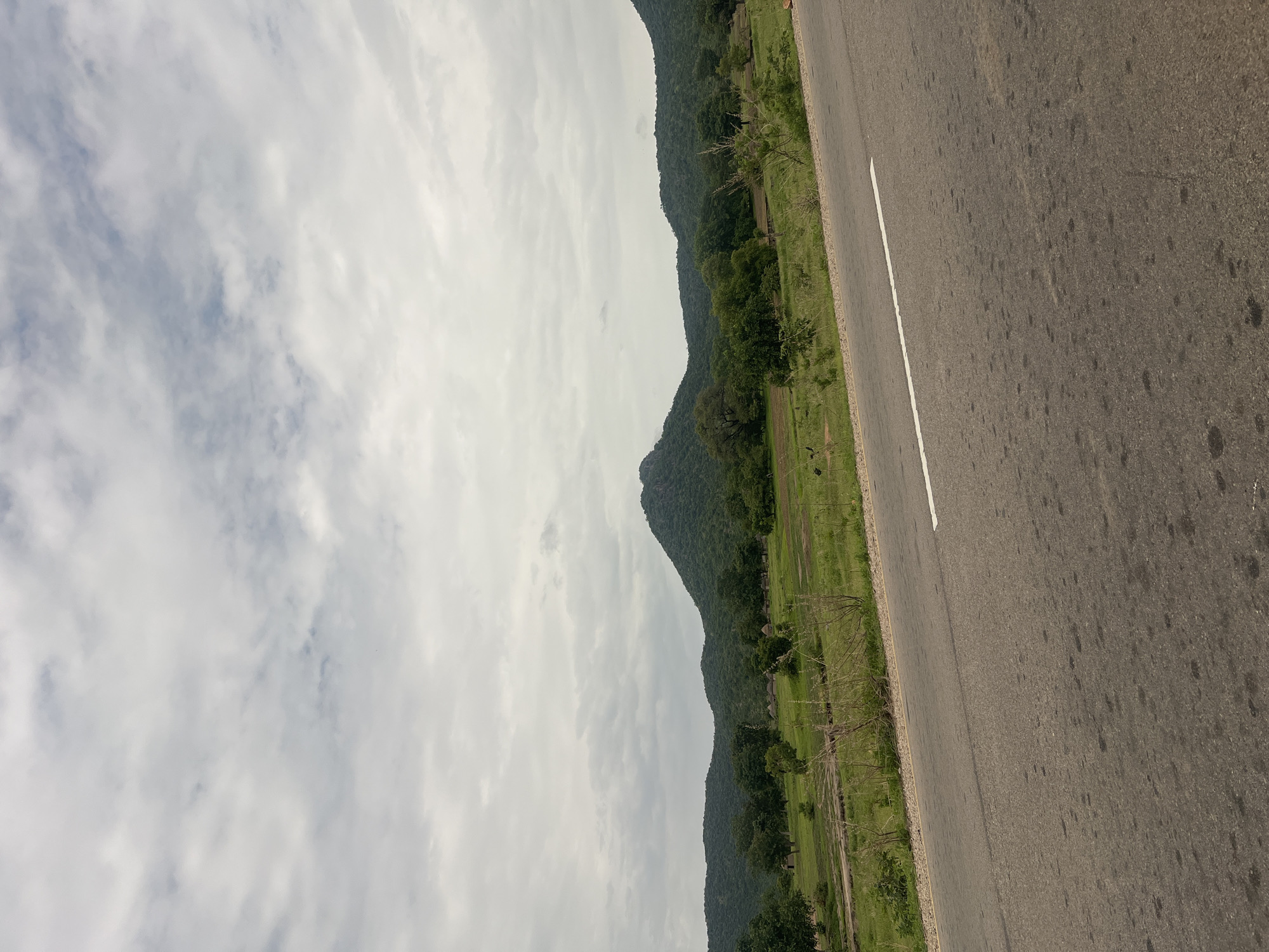 This is a vertical-oriented photograph of a paved road running alongside a lush, green landscape and a series of undulating hills or mountains covered in dense trees. The road appears to be a two-lane highway with a white line marking the center. The right side of the road is asphalt, showing some dark splotches. The left side of the road is bordered by a grassy verge with some scattered trees and vegetation.
Beyond the roadside greenery, the land stretches out, appearing to be fields or open grassy areas, interspersed with more trees and patches of darker green, possibly indicating denser foliage or cultivated areas. The background is dominated by a chain of low, wooded hills that recede into the distance. The sky is overcast with scattered clouds, suggesting a potentially dull or humid weather condition. The lighting is diffused, implying it is daytime, but the exact time is hard to determine due to the cloud cover.
There are no people or distinct objects visible in the scene, emphasizing a natural landscape. The overall impression is one of a rural or semi-rural setting, likely in a tropical or subtropical region given the verdant vegetation. The location context provided is Minda, Nigeria, which aligns with the visual cues of a tropical environment and the type of road construction. The image conveys a sense of peacefulness and natural beauty. No text is visible in the photograph.