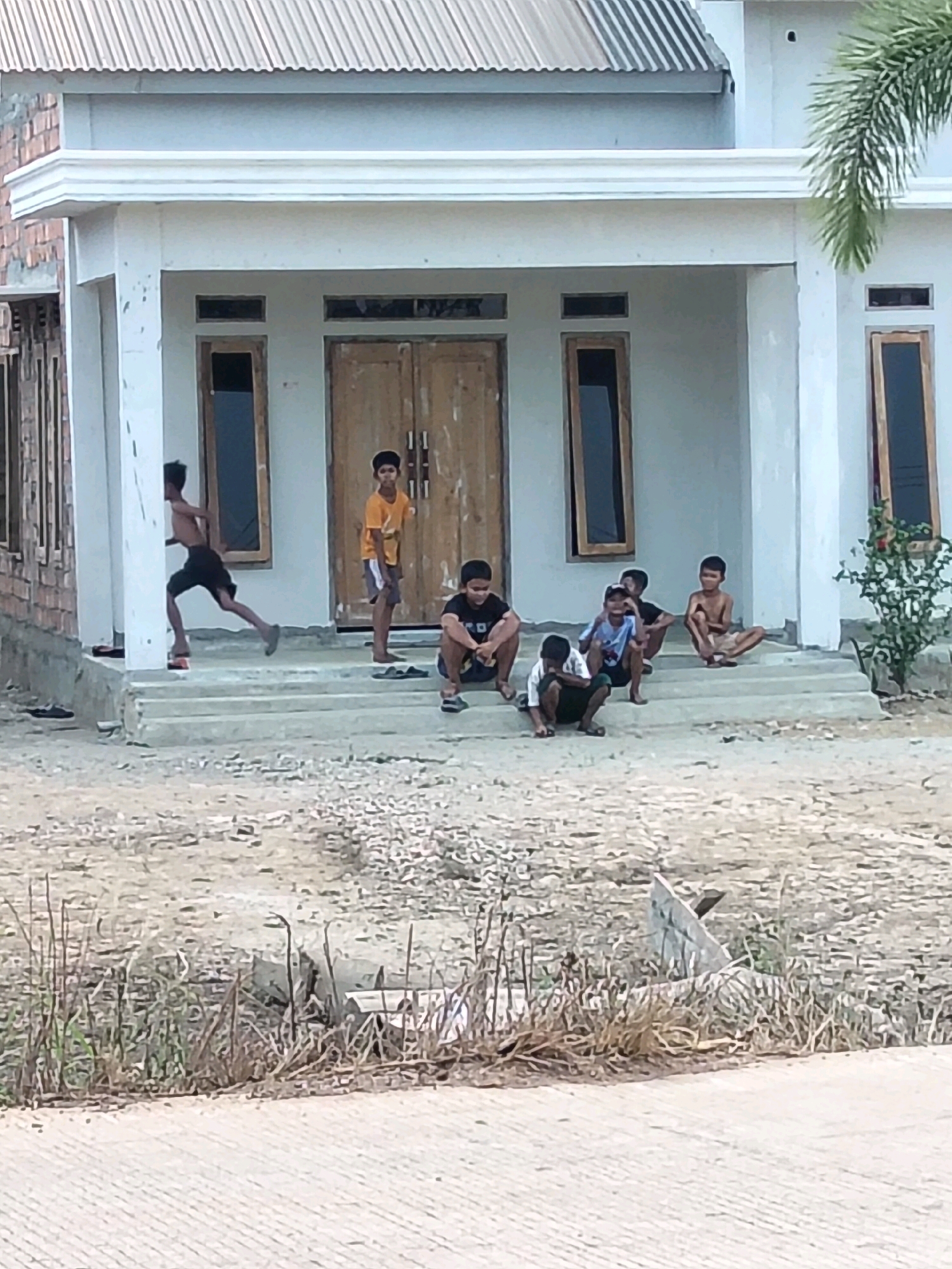 The image depicts a group of seven young boys in Perambahan, Indonesia, on the steps and front yard of a house. The house has a white facade, a corrugated metal roof, and wooden double doors. The boys are casually dressed, and their activities suggest a relaxed, informal gathering.
On the steps, from left to right:
* A boy in black shorts is running on the second step from the bottom, appearing to be in motion.
* A boy in a yellow t-shirt and grey shorts stands on the third step from the bottom.
* A boy in a black t-shirt and dark shorts is seated on the fourth step from the bottom.
* A boy in a blue t-shirt and black shorts is crouched on the fourth step from the bottom.
* A boy with a bare chest and light-colored shorts is seated on the fourth step from the bottom.
In front of the steps, on the ground:
* A boy wearing a white t-shirt and dark shorts is crouched on the ground.
* A boy in a black t-shirt and dark shorts is also crouched on the ground.
The environment surrounding the house is characterized by dry, rough terrain with sparse vegetation, including dried grasses and some small bushes. The foreground shows a paved surface, likely a road or pathway, with a textured pattern. The overall scene conveys a sense of everyday life in a residential area in Indonesia.