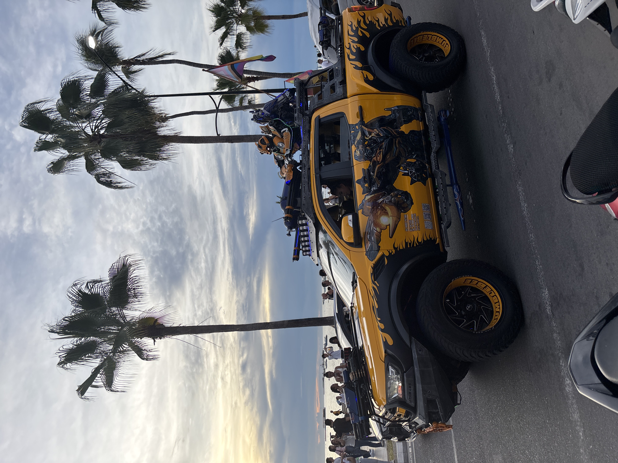 This image depicts a highly customized yellow Ford Ranger pickup truck, likely a prop from a Transformers movie, on a street in Pattaya City, Thailand. The truck features intricate graphics of Bumblebee, the Transformers character, along its sides and is equipped with a roof rack holding what appear to be replica weapons and robot figures. A rainbow pride flag is also visible, suggesting it might be part of a parade or event. The truck is adorned with flame decals and has modified tires with the word "BUMBLEBEE" on the rims. In the background, palm trees line the street, and a body of water, presumably the Gulf of Thailand, is visible, with a hazy sky indicating it is likely sunset or early evening. Several people are gathered on the sidewalk, observing the decorated vehicle. The overall scene suggests a festive or promotional event taking place in a coastal urban environment.