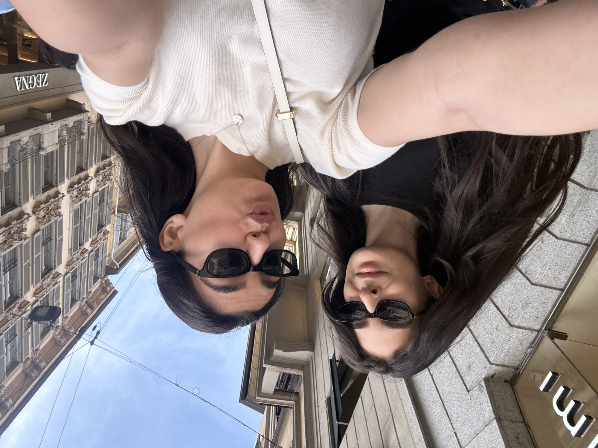 Two women in sunglasses take a selfie while standing on a street in Milan, Italy. One woman is wearing a white shirt and the other is wearing a black shirt. The Zegna store is visible in the background. The other discernible brand is the Loro Piana store. The architectural style of the building also suggests a European location.
