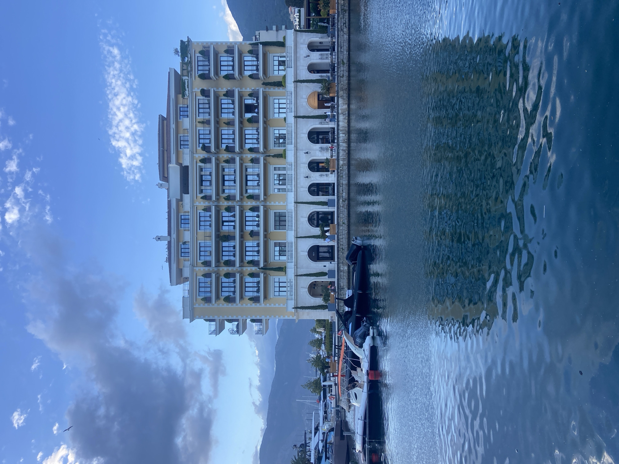 The image depicts a multi-story building with a yellow and white facade located in Tivat, Montenegro. The building has several balconies and windows. The building faces a body of water, possibly a harbor or marina, where boats and yachts are docked. The reflection of the building can be seen in the water. In the background, mountains are visible. The sky above contains scattered clouds.