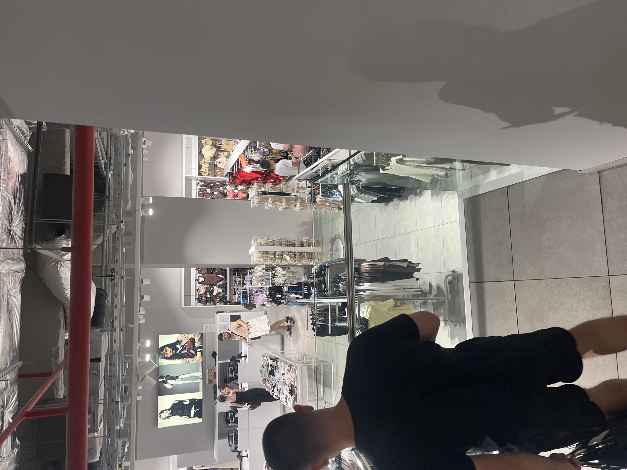 The image depicts the interior of a retail store, likely a clothing store. The location is unspecified and there is no indication that the image was taken in Blagoevgrad, Bulgaria. Clothing racks and displays are visible, stocked with merchandise. Several people are present in the image. A man wearing black shorts and a black t-shirt is standing in the foreground, partially obscuring the view. A man wearing a white outfit can be seen in the middle ground. A woman wearing black is standing behind a counter. The lighting appears to be fluorescent, and the environment is commercial.