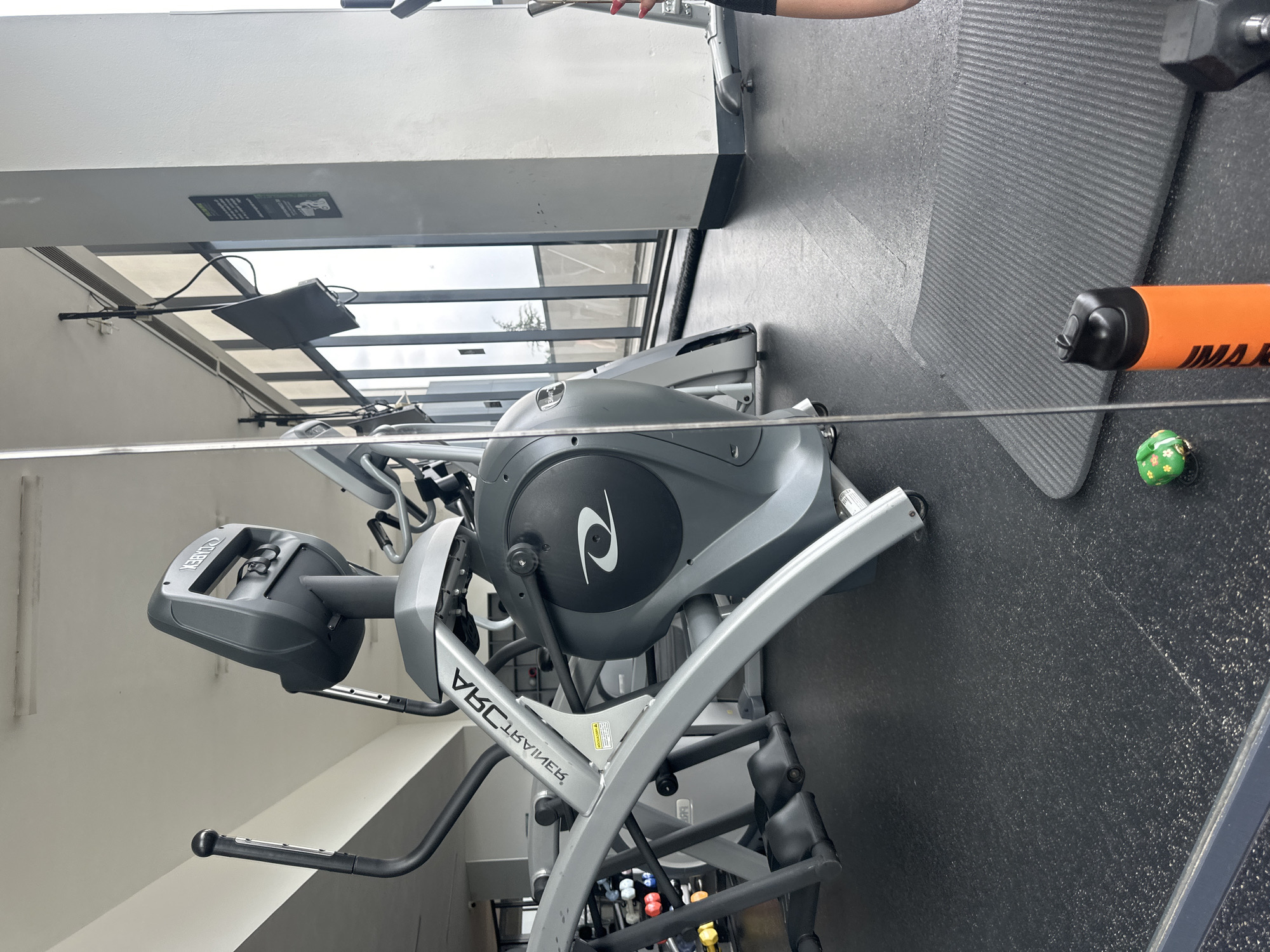 The image depicts a gym setting in Huixquilucan de Degollado, Mexico. An elliptical machine dominates the foreground. A black rubber mat is visible to the right of the elliptical. An orange water bottle and a small green decorative ball are present on the mat. Gym windows and structural columns are visible in the background.