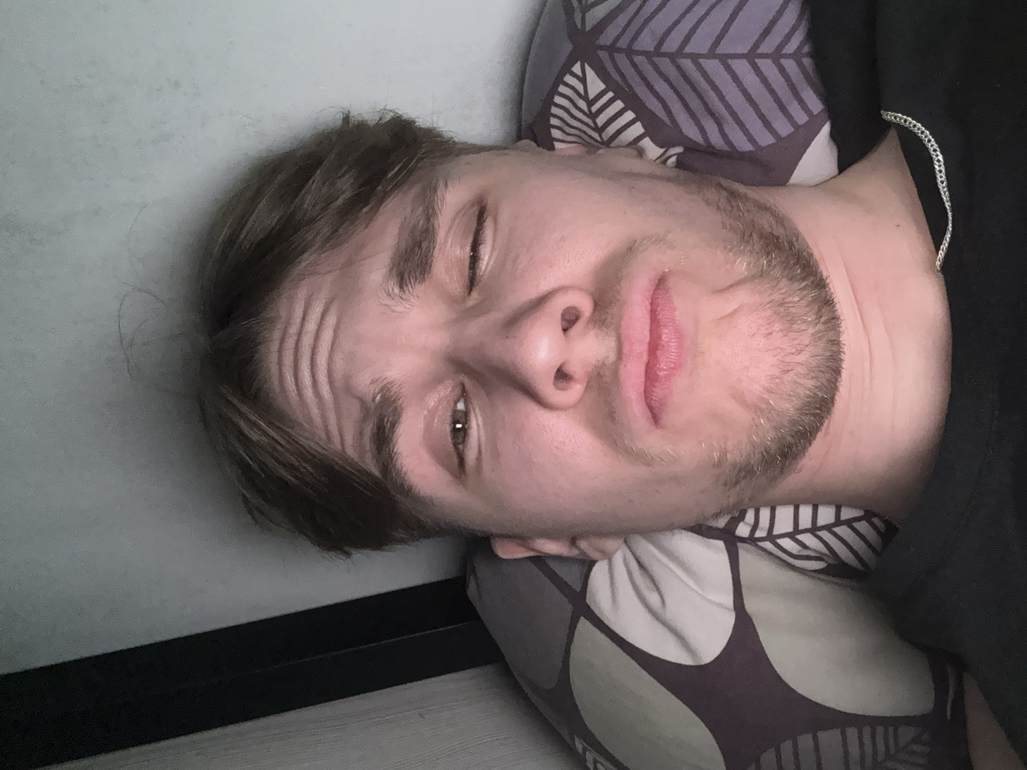 A man lies on a pillow, his head tilted, with one eye closed and a slight frown. He has short brown hair, stubble, and visible forehead wrinkles. He appears to be indoors, possibly on a bed or resting against a wall. The surroundings are indistinct but suggest a domestic setting.