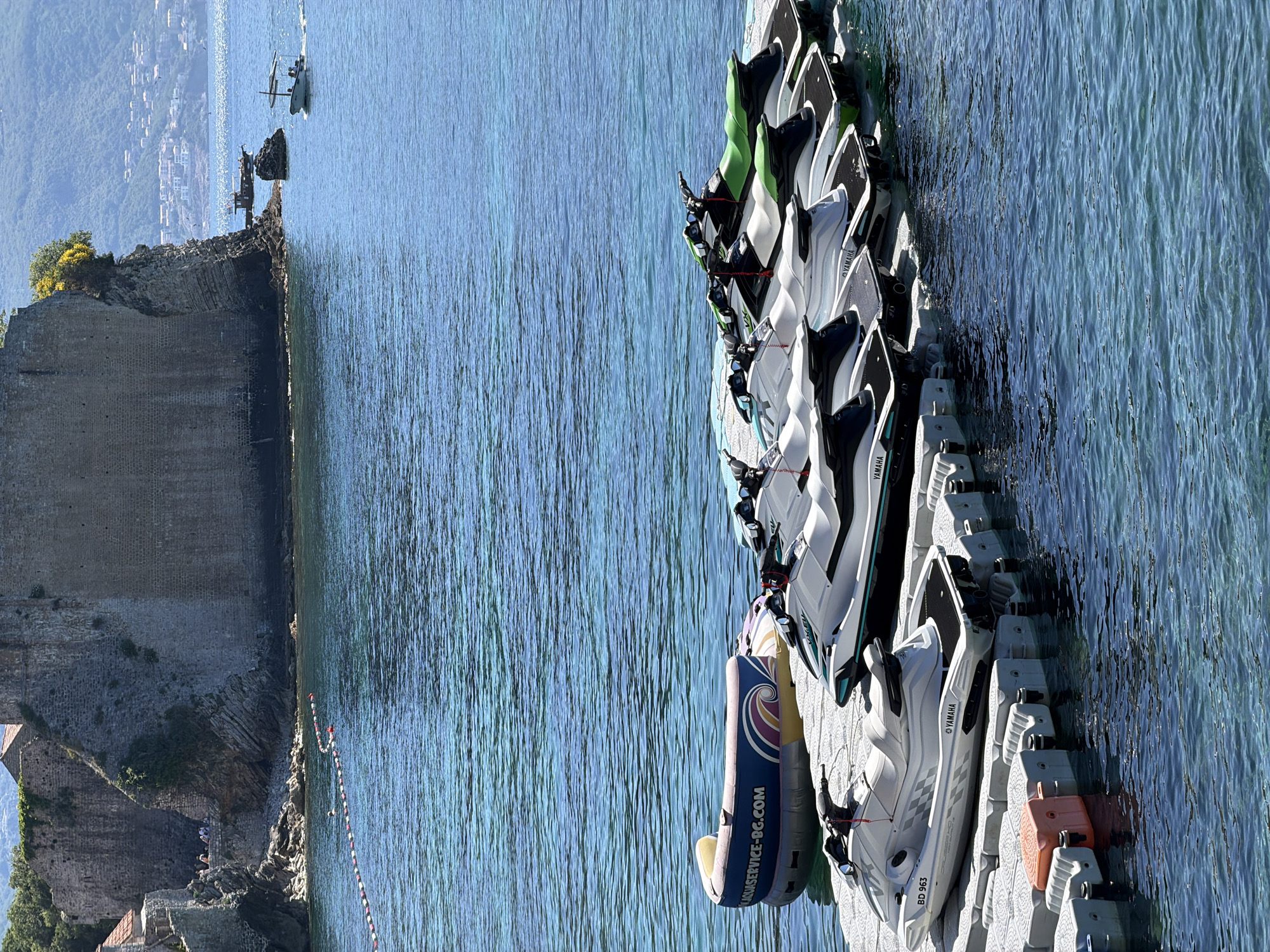 The image shows a view in Budva, Montenegro, featuring a stone structure, likely part of the old city walls or a fortress, on the left. To the right is the sea, with a floating dock holding a row of jet skis, predominantly Yamaha models. Some inflatable tubes are also present. A boat can be seen in the distance. The sea is relatively calm.