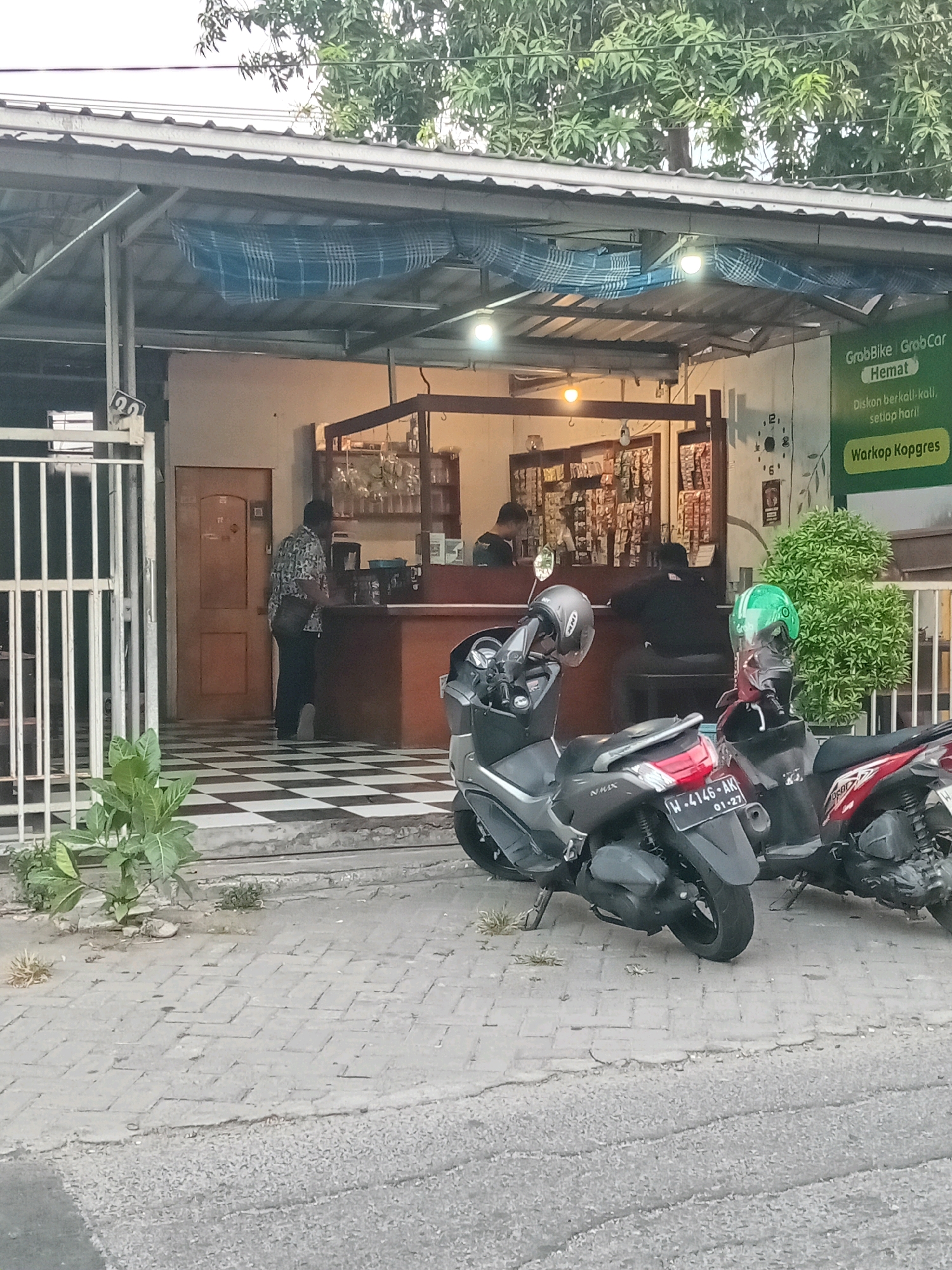 The image shows the exterior of a small shop or eatery, identified by a sign as "Warkop Kopgres," located in Randuagung, Indonesia. The shop has a covered outdoor seating area with a checkered tile floor. In front of the shop, there are two motorcycles parked on a paved sidewalk. One motorcycle is gray and black, and the other is red and black. The gray motorcycle has a license plate reading "W-4146-AK 01-27". Helmets are visible on the handlebars of both motorcycles. Inside the shop, shelving units are filled with products, and a counter is visible. Several individuals are present inside the shop, with one person standing behind the counter and another seated. The shop appears to be open for business, as indicated by the lighting and activity. The overall environment suggests a typical urban or semi-urban street scene in Indonesia, with a focus on the local business.