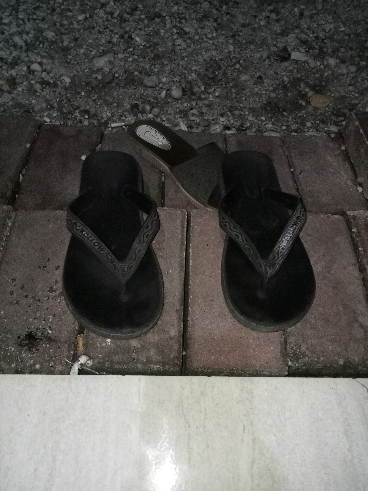 In the city of Kualu, Indonesia, on a paved walkway with a gravel-covered area behind it, two pairs of black flip-flops and one pair of black slide sandals are displayed. The flip-flops are positioned side-by-side, with the slide sandals placed slightly behind them. The lighting in the scene is dim, suggesting it is either nighttime or a dimly lit outdoor space. The walkway is composed of rectangular pavers, and the gravel area contains small stones and pebbles. A few cigarette butts are scattered on the ground near the flip-flops. The overall impression is one of casual outdoor footwear placed on a rough, utilitarian surface.