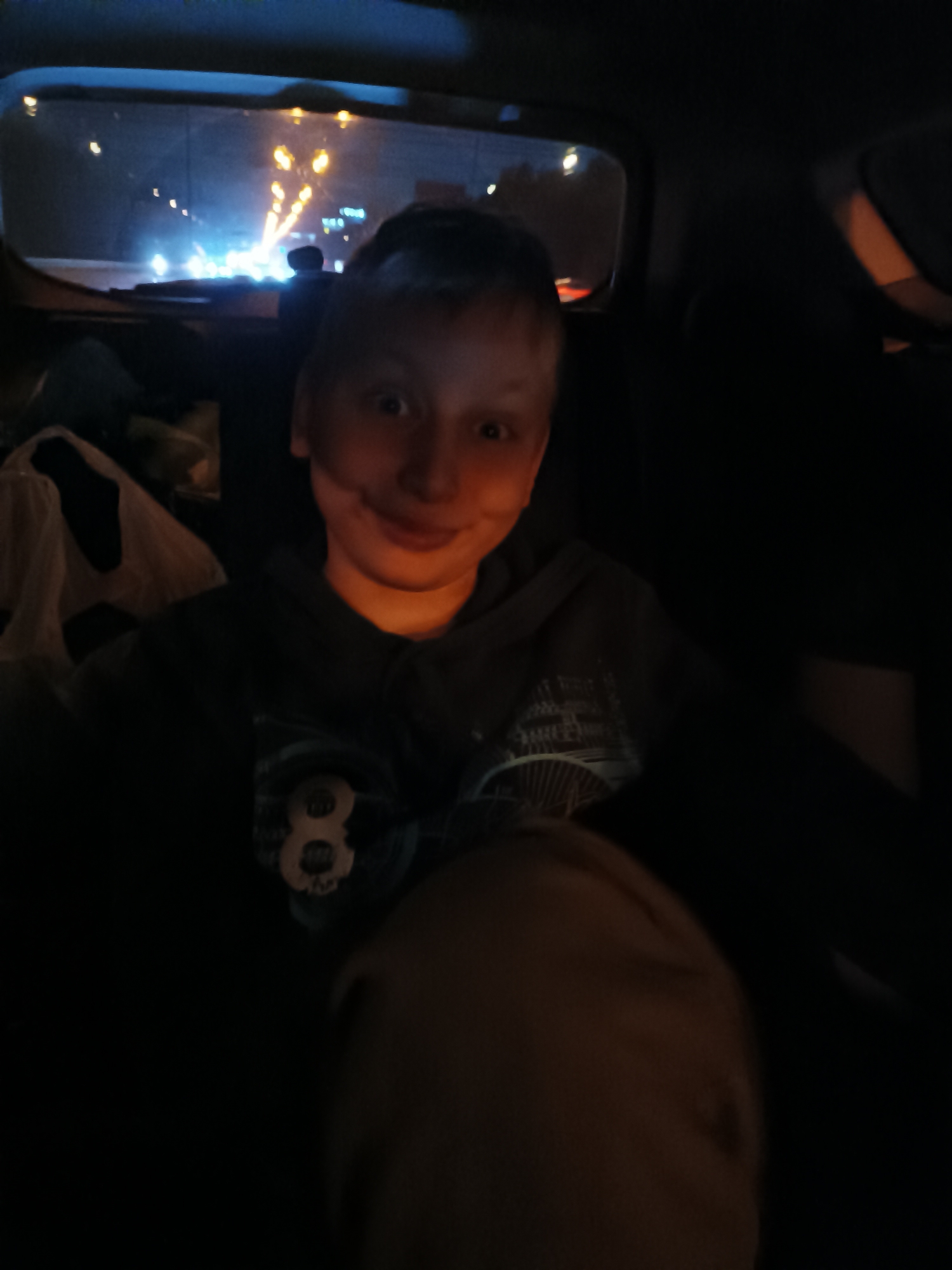 In the low-light interior of a vehicle, a young person with fair skin and short, dark hair is looking at the camera with a slight smile. They are wearing a dark hooded sweatshirt with a graphic design on the chest, featuring a prominent number "8". The background shows a nighttime view through the rear window, illuminated by streetlights and the headlights of other vehicles, suggesting an urban environment. The scene is consistent with being in a car at night in a city such as Moscow, Russia, given the typical urban lighting and vehicle presence.