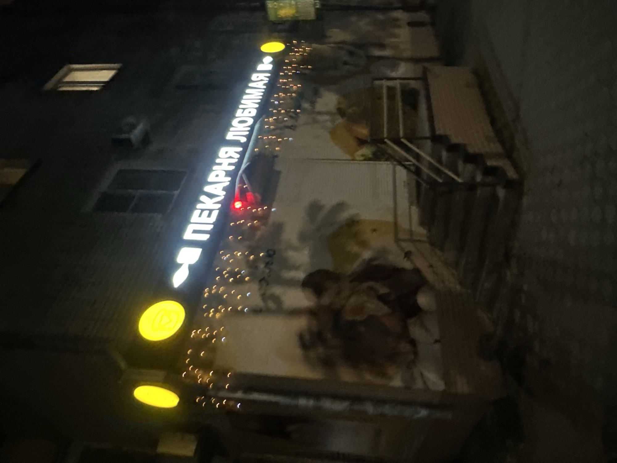 The image depicts the exterior of a building at night, likely a commercial establishment, in Birobidzhan, Russia. The primary subject is a vertical illuminated sign in Cyrillic script that reads "ПЕКАРНЯ ЛЮБИМАЯ," which translates to "Beloved Bakery." The sign is white and outlined, with a stylized flame symbol preceding the text. Below the main sign, two large circular yellow lights are visible, one above the other. A string of small, warm-toned lights is draped along the building facade, adding to the festive or inviting atmosphere.
The building appears to be multi-story, with at least one visible window illuminated from within. The overall setting is dark, consistent with nighttime. There are some architectural elements visible, including what looks like a staircase to the right of the sign, and some indistinct shapes that might be people or objects obscured by the low light and low resolution. The texture of the building's facade appears rough. The scene evokes a sense of a neighborhood bakery operating in the evening or at night.
