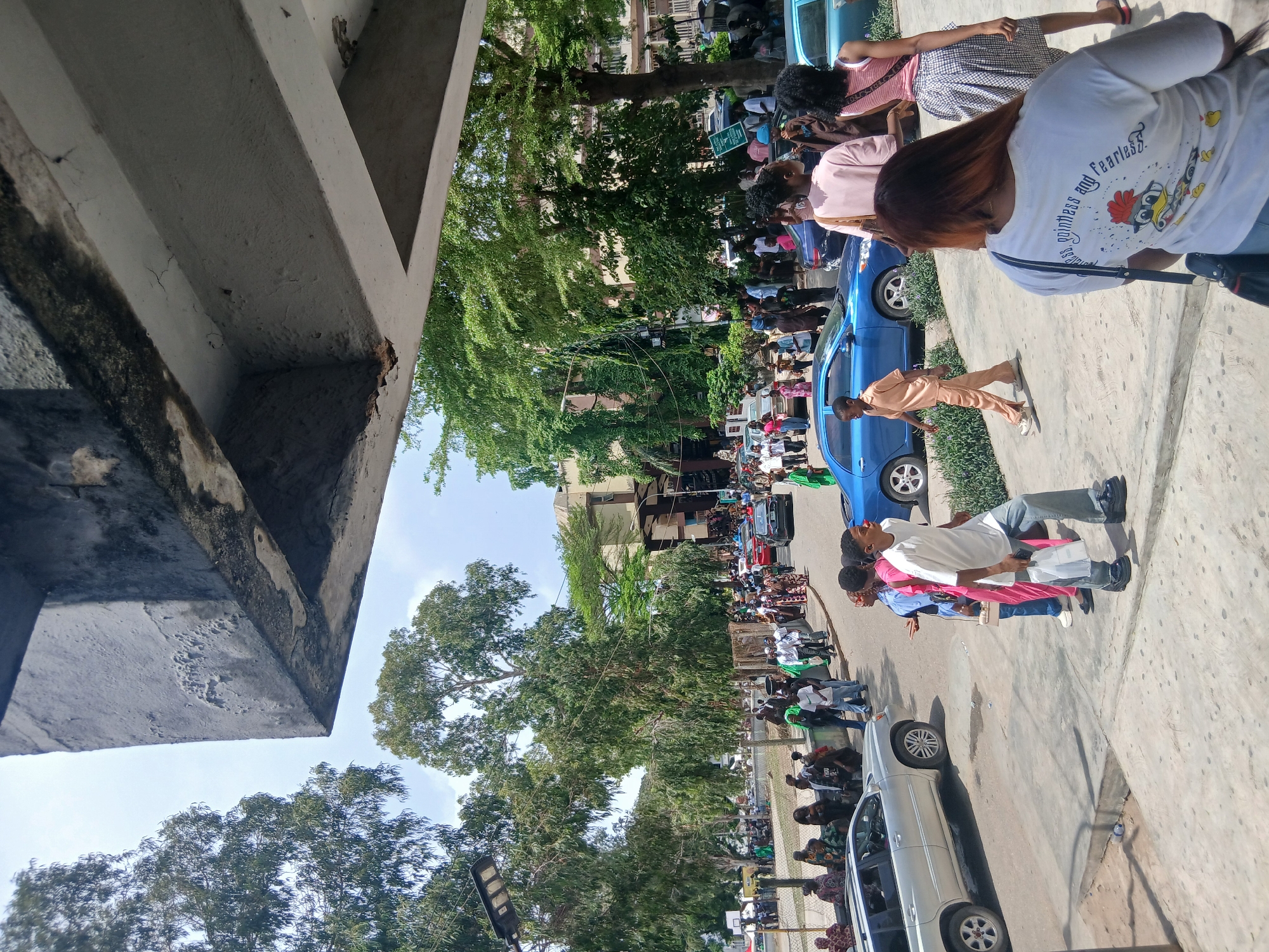 A daytime, street-level view showing a crowd of people gathered along a sidewalk in what appears to be a demonstration or public gathering in Lagos, Nigeria. A concrete structure is visible on the left side of the frame. Cars are parked along the street, including a silver SUV, blue sedan, and possibly other vehicles obscured by the crowd. The crowd consists of many individuals of diverse appearances. There are green trees visible behind the crowd and buildings, possibly apartments, visible in the background. There is a light blue sky with some clouds.