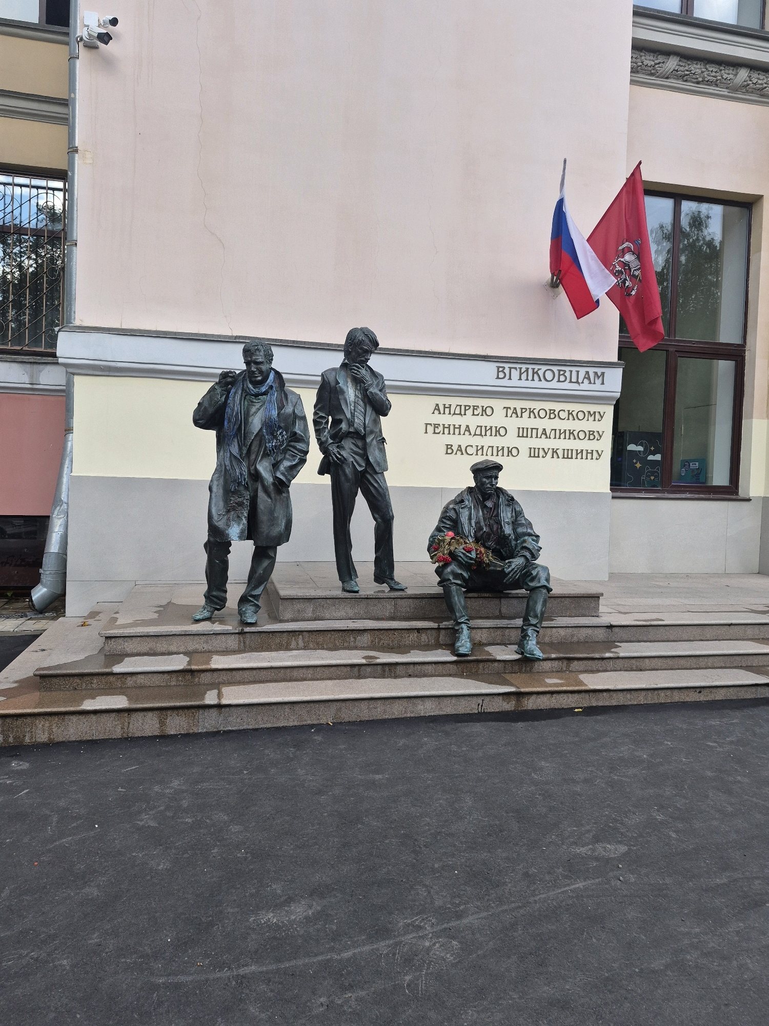 The image shows a bronze sculpture of three men standing and sitting on a set of stone steps in front of a building. The man on the left stands with his hand in his pocket and a scarf around his neck. The man in the middle stands with his hand touching his face, looking down. The man on the right sits on the steps, holding a bouquet of flowers. Above the sculptures, text is visible on the building wall, and two flags are mounted on the wall. The Russian flag and a red flag with a coat of arms are displayed. The building appears to be a public institution, possibly an educational facility, given the presence of security cameras mounted on the exterior wall. The ground in front of the steps is paved and appears wet, suggesting recent rain. The scene is set outdoors, with the building and statues illuminated by daylight. This monument is located on a side street in Moscow, Russia, specifically on the corner of Mytnaya Street and the small lane "Malaya Mytnaya" where the VGIK film institute is located. The monument honors Andrei Tarkovsky, Gennady Shpalikov, and Vasily Shukshin, who were prominent figures in Soviet cinema.