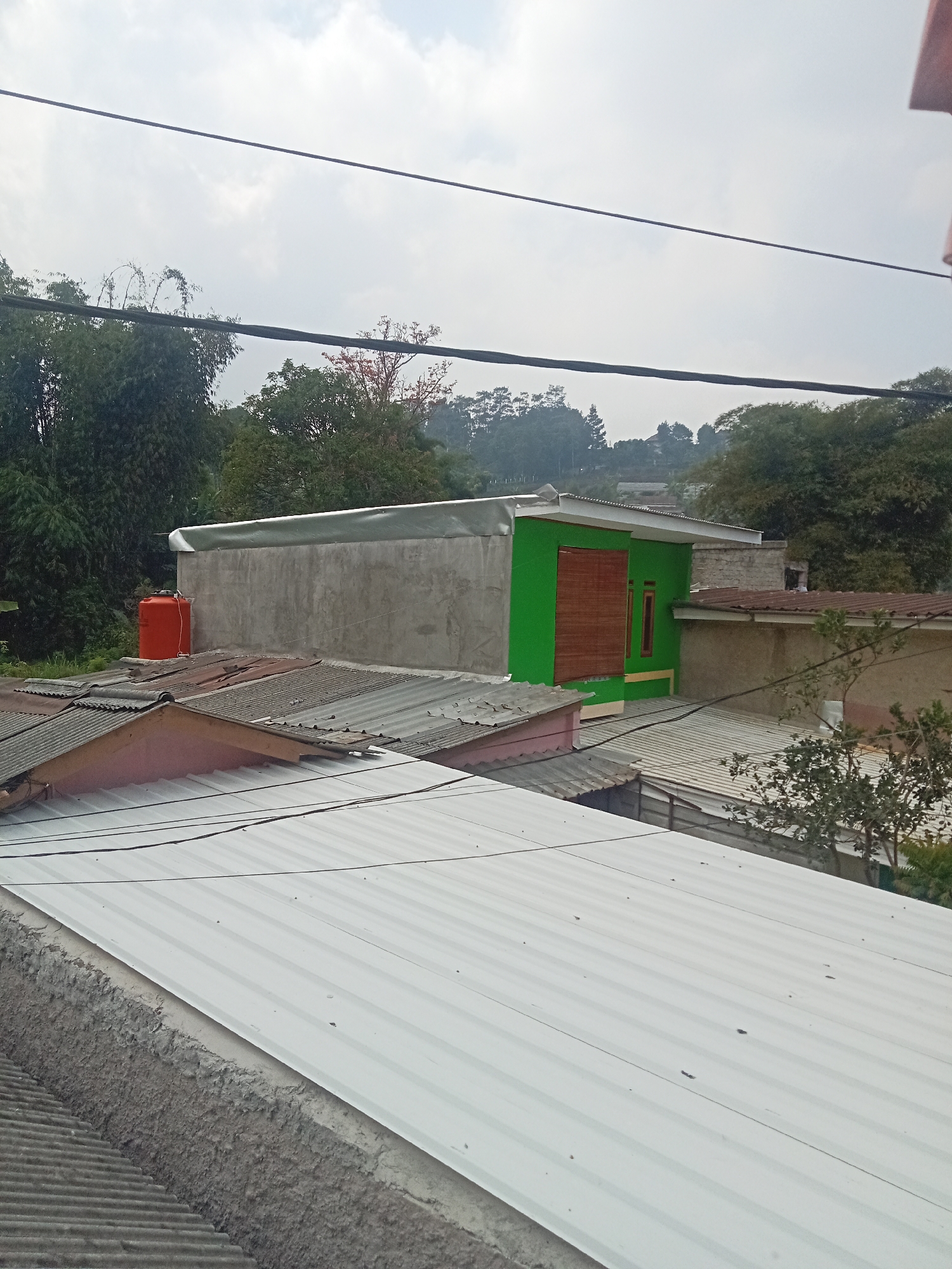 The image captures a rooftop view in Pagerwangi, Indonesia, dominated by the corrugated metal roofs of several buildings. In the foreground, a large, white corrugated metal roof occupies the majority of the frame, with its textured surface showing subtle lines and a slightly uneven finish. To the left and slightly behind this prominent roof, another corrugated metal roof, this one dark gray and ribbed, is visible. Further into the mid-ground, a pink-colored roof segment with some visible damage or wear is present.
Behind these lower structures, a more substantial building is partially visible. It features a concrete wall and a prominent section painted a vibrant green. This green section has a wooden-slatted window covering and a smaller, dark-framed window. A bright orange water tank is positioned on a roof adjacent to the concrete wall. The background is filled with lush green trees and vegetation, suggesting a semi-urban or rural setting within the city. The sky above is overcast, with visible power lines stretching across the upper portion of the image. No people are visible in the frame.