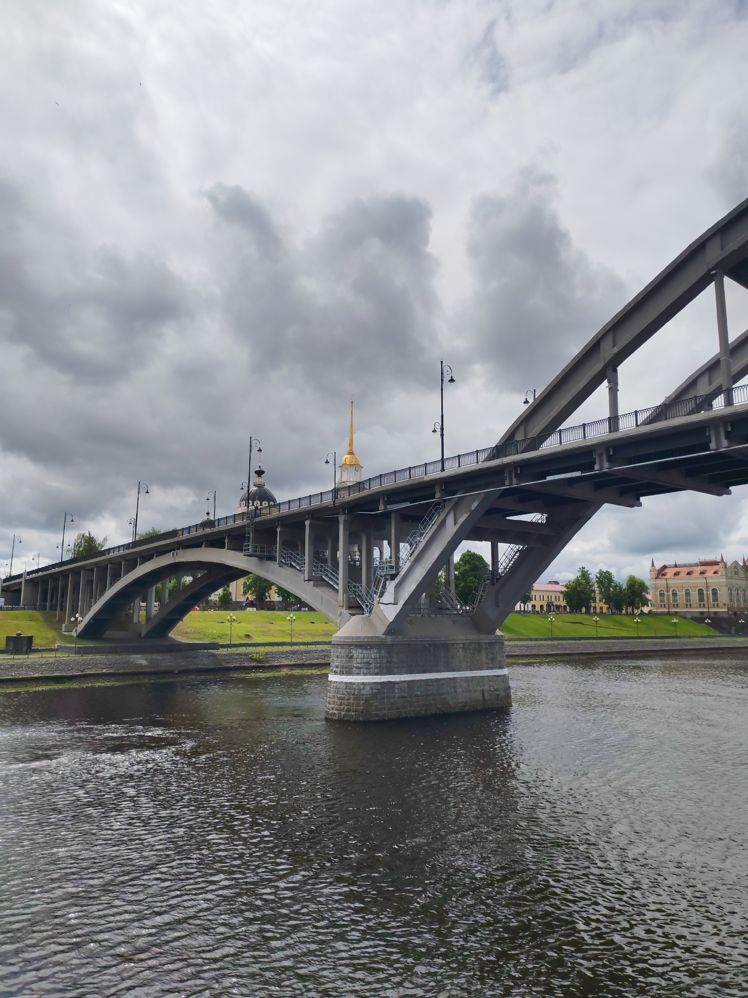 The image depicts a bridge in Rybinsk, Russia, under an overcast sky. The bridge is constructed of concrete and steel, featuring both arched and truss sections. It spans a body of water with ripples visible on the surface. On the banks are green grassy areas and buildings, one with a prominent gold spire. Lampposts line the bridge's length. No people are visible in the image.