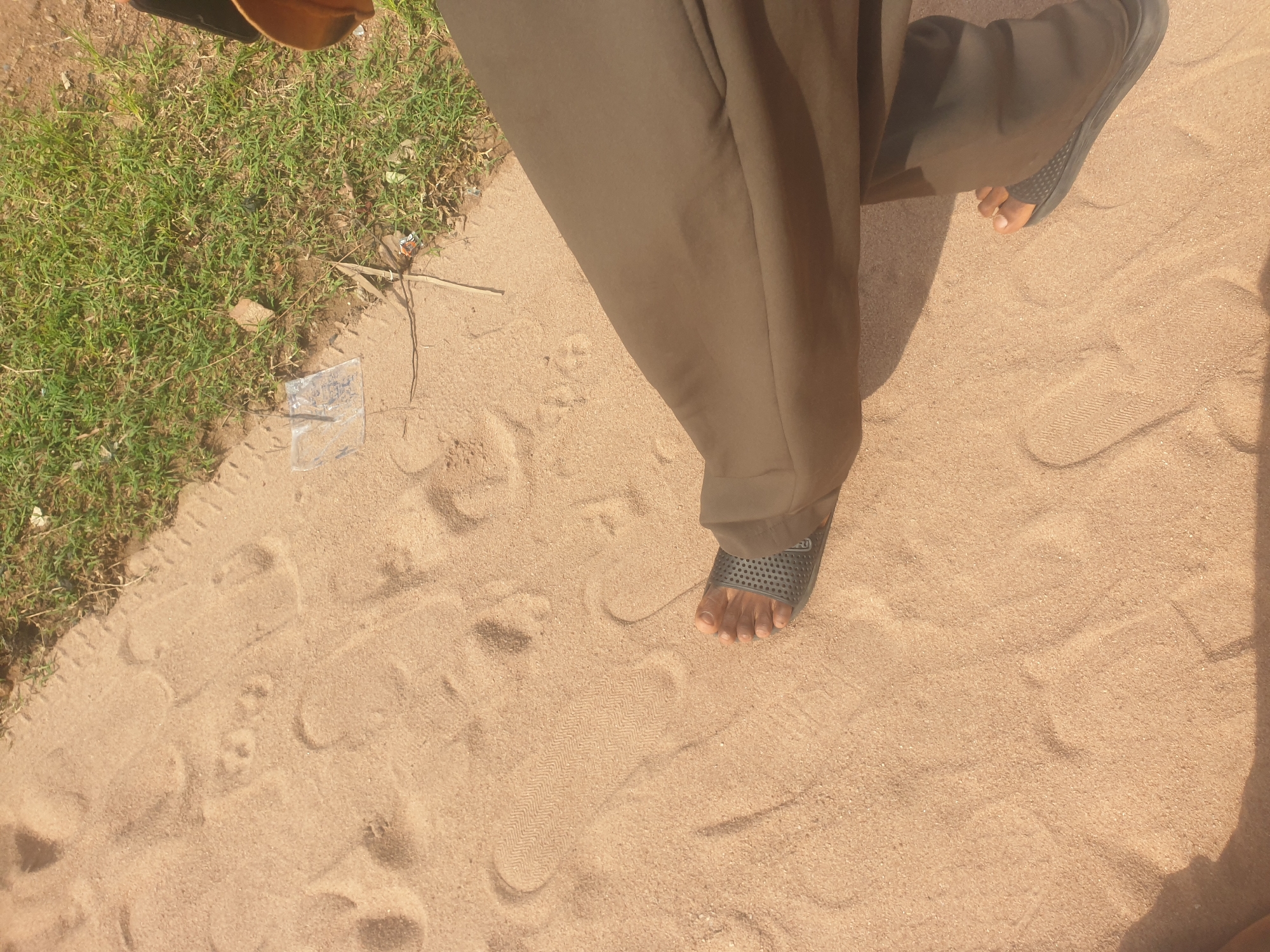 The image shows a person's feet and legs standing on a sandy surface next to a patch of grass. The sand is light brown and has several footprints impressed in it. The grass is green and appears to be growing on a slightly elevated area next to the sand. There is a small piece of clear plastic trash on the sand near the grass. The person is wearing dark brown pants and black sandals. The scene is likely outdoors in a natural setting.