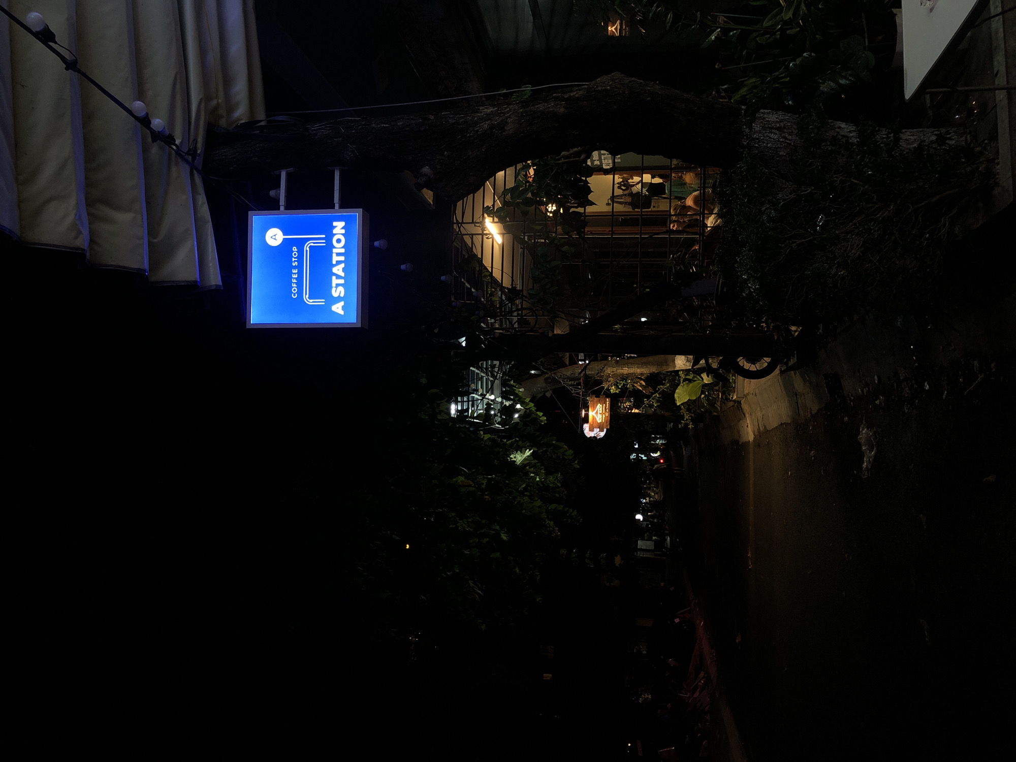The image depicts a nighttime scene in what appears to be an outdoor cafe or restaurant setting in Da Nang, Vietnam. The primary subject is a brightly lit blue sign that reads "A STATION COFFEE STOP" in white text. The sign is suspended from a metal rod, and several string lights with exposed bulbs hang above it, casting a warm glow.
Behind the sign, a large, gnarled tree trunk is prominently featured, arching over a windowed area. Through the window, interior lights illuminate the space, revealing glimpses of people seated inside. The overall atmosphere suggests a cozy and inviting establishment, with the contrasting dark exterior and warm interior lighting creating a sense of welcome.
Foliage is visible throughout the scene, suggesting a natural and possibly overgrown environment. The time of day is clearly night, indicated by the darkness and the artificial lighting. Location cues, such as the style of the signage and the outdoor setting, are consistent with a cafe in Vietnam. No specific weather conditions are discernible, but the scene is generally dark and illuminated by artificial light sources. The overall emotion evoked is one of calm and possibly relaxation, characteristic of a coffee shop setting at night.