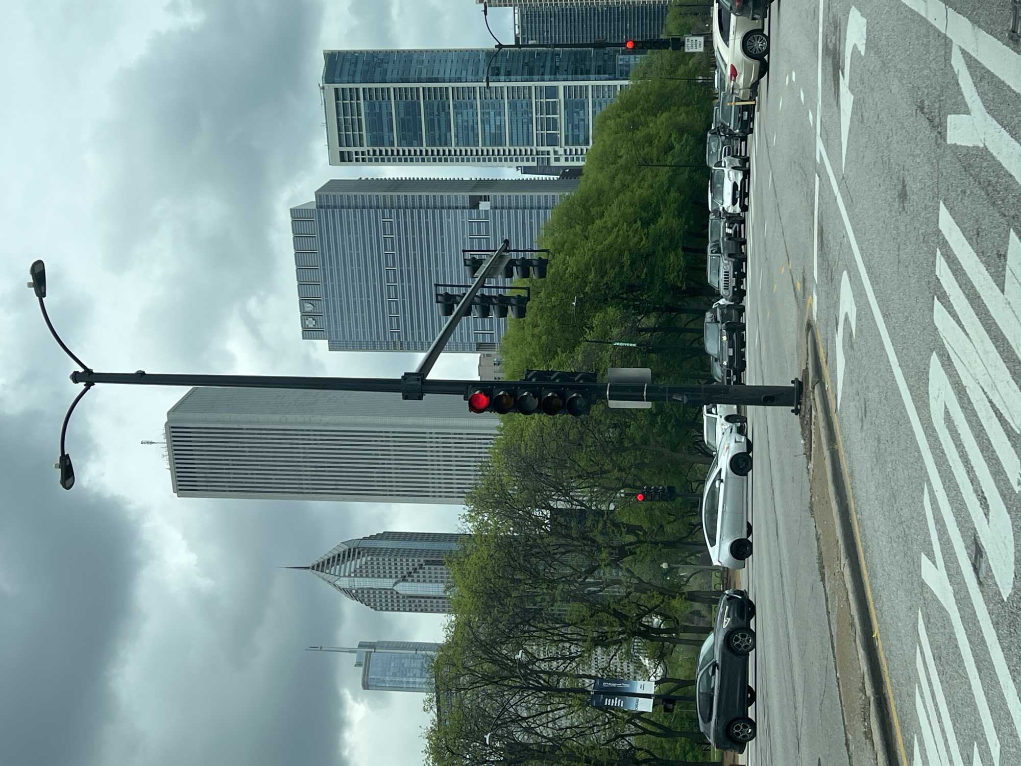 The image captures a street scene in Chicago, United States of America, under an overcast sky. Several skyscrapers dominate the background, their architectural details only partially visible through the low-hanging clouds. A row of green trees lines the left side of the street, adding a touch of nature to the urban landscape.
On the right side of the frame, cars are lined up along the curb, and a crosswalk with painted white stripes is visible. Traffic lights are strategically placed on black poles, with at least one showing a red light. A street light with a curved arm extends from a pole on the left.
