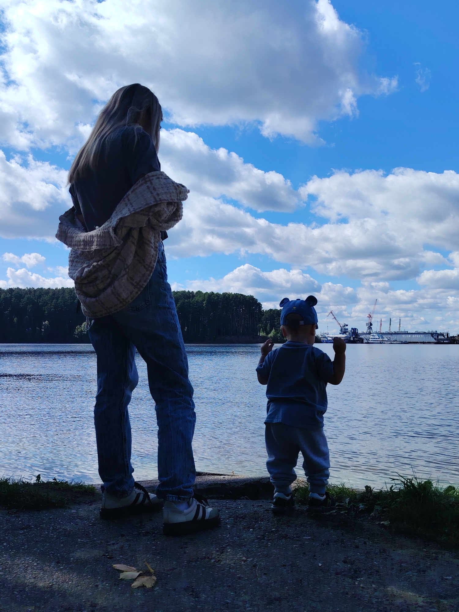 An adult and a child stand on the shore of a body of water, looking out at a harbor with cranes and ships. The adult, seen from behind, has long blonde hair and is wearing a dark shirt with a patterned scarf draped over their shoulders and blue jeans. The child, also seen from behind, wears a blue shirt, grey pants, and a blue hat with bear ears. The water is calm, reflecting the blue sky dotted with white clouds. A dense forest lines the opposite shore, and the harbor is in the distance. The ground in the foreground is a mixture of dark gravel and sparse grass. The image appears to have been taken on a sunny day. This scene is likely the Chaikovsky Reservoir in Chaikovsky, Russia, a popular recreation area and a key feature of the city's landscape, often used for various water activities and known for its scenic views of the Kama River.