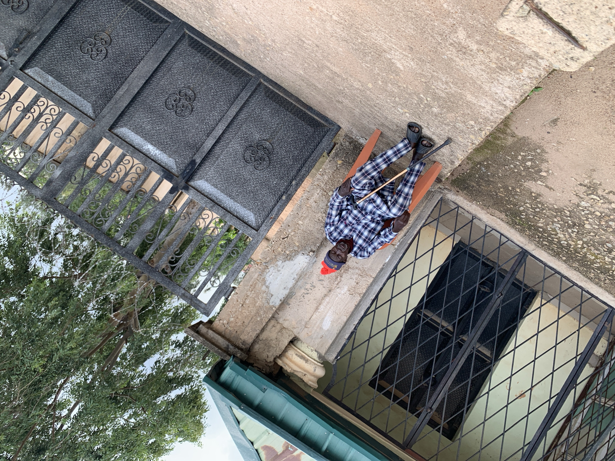 In Jos, Nigeria, an elderly Black man in a blue and white plaid outfit and a red beanie sits on an orange plastic chair outdoors. He holds a wooden walking stick in his right hand. The man is positioned next to a building with a green corrugated metal roof and a barred window. Behind him is a large, dark metal gate with an ornate design. The scene appears to be a residential area, with concrete ground and a tree visible in the background.