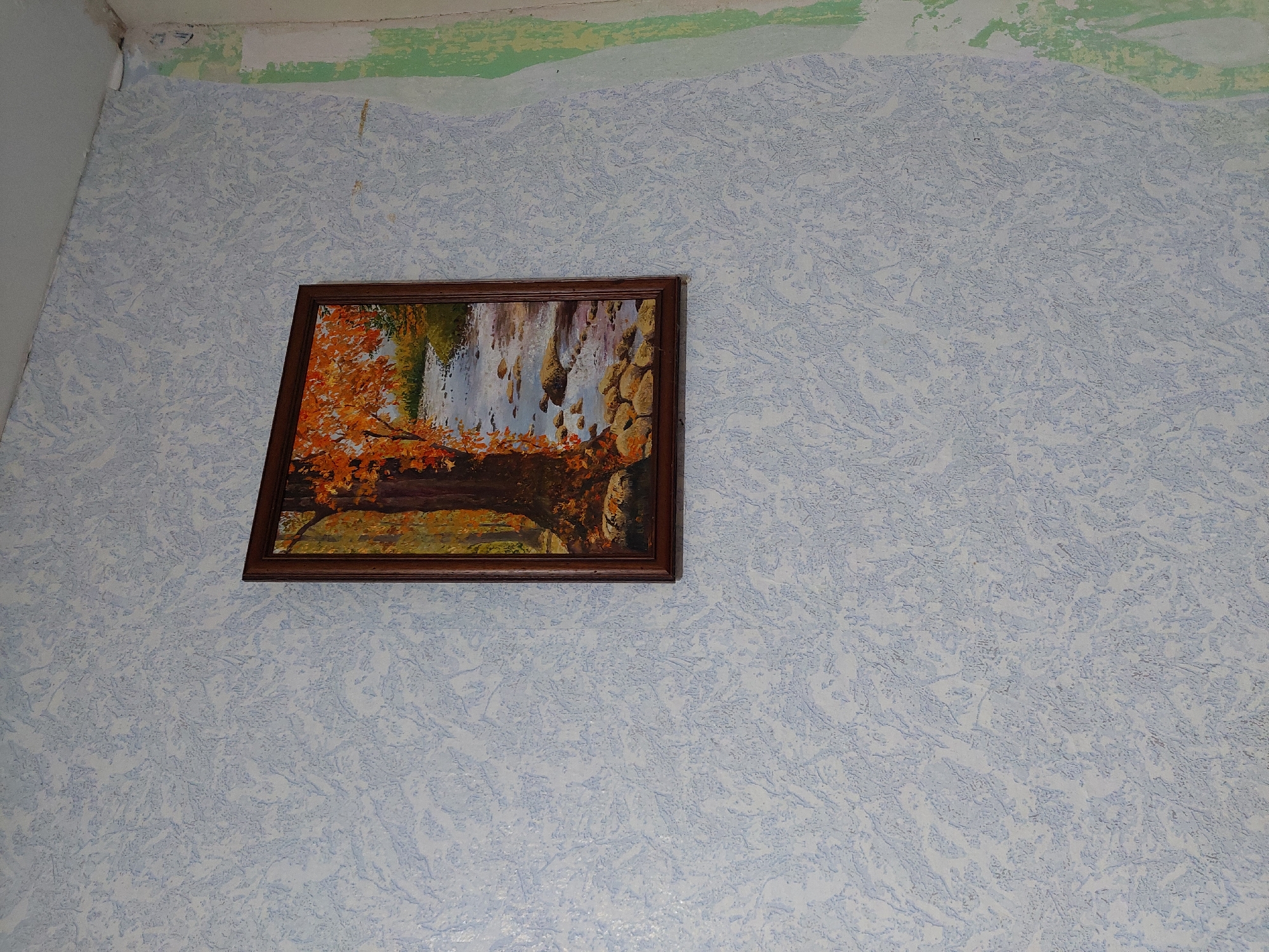 An indoor scene in Orhei, Moldova, depicts a framed painting affixed to a textured wall. The painting, oriented vertically, features an autumnal landscape with a large tree exhibiting orange foliage on the left, beside a rocky river or stream flowing towards the background. The frame is a dark, possibly wooden, rectangular structure. The wall is covered in light blue wallpaper with a mottled, abstract, or stippled pattern. To the right, the wall transitions into an adjacent surface or corner where the wallpaper is absent, revealing layers of light green and lighter-toned material, indicating a state of partial renovation or disrepair. No individuals are present, and no actions are discernible.