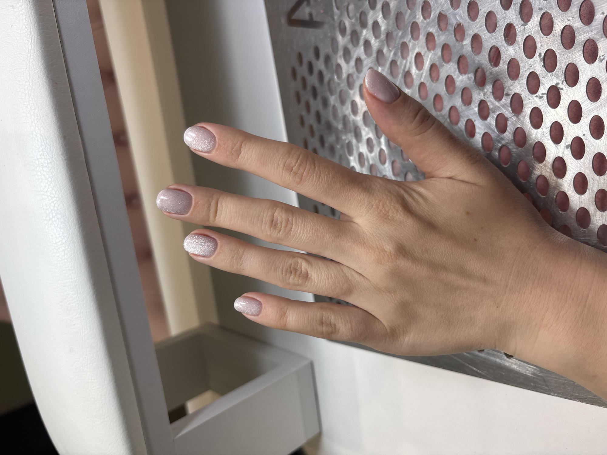 A medium shot captures a fair-skinned hand with professionally manicured nails. The nails are painted in a light, neutral color with a subtle, glittery effect. The hand is positioned against a perforated metal surface. A white piece of furniture or equipment is visible in the background.
The location in the city of Ферма, Russia is not discernible from the image.