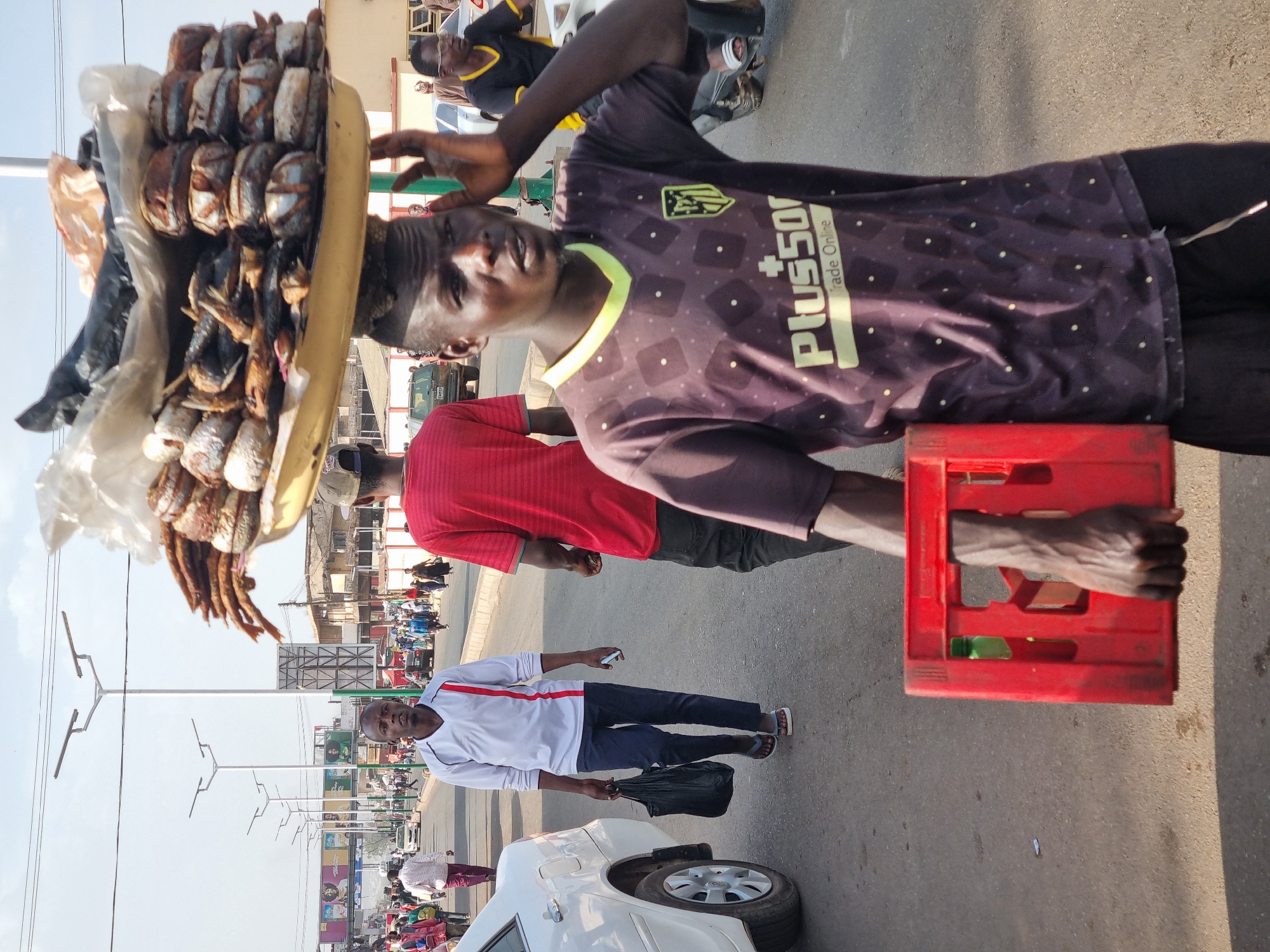 A street scene in Jos, Nigeria, depicts a vendor wearing a dark t-shirt with a soccer club logo and the "Plus500" sponsorship emblem. He carries a red crate in one hand and a tray balanced on his head filled with what appears to be smoked fish. Several other people populate the background, including a man in a white shirt with a red stripe and a black bag. Cars are parked along the side of the road, and street lights are visible. Other shops are observed in the background.